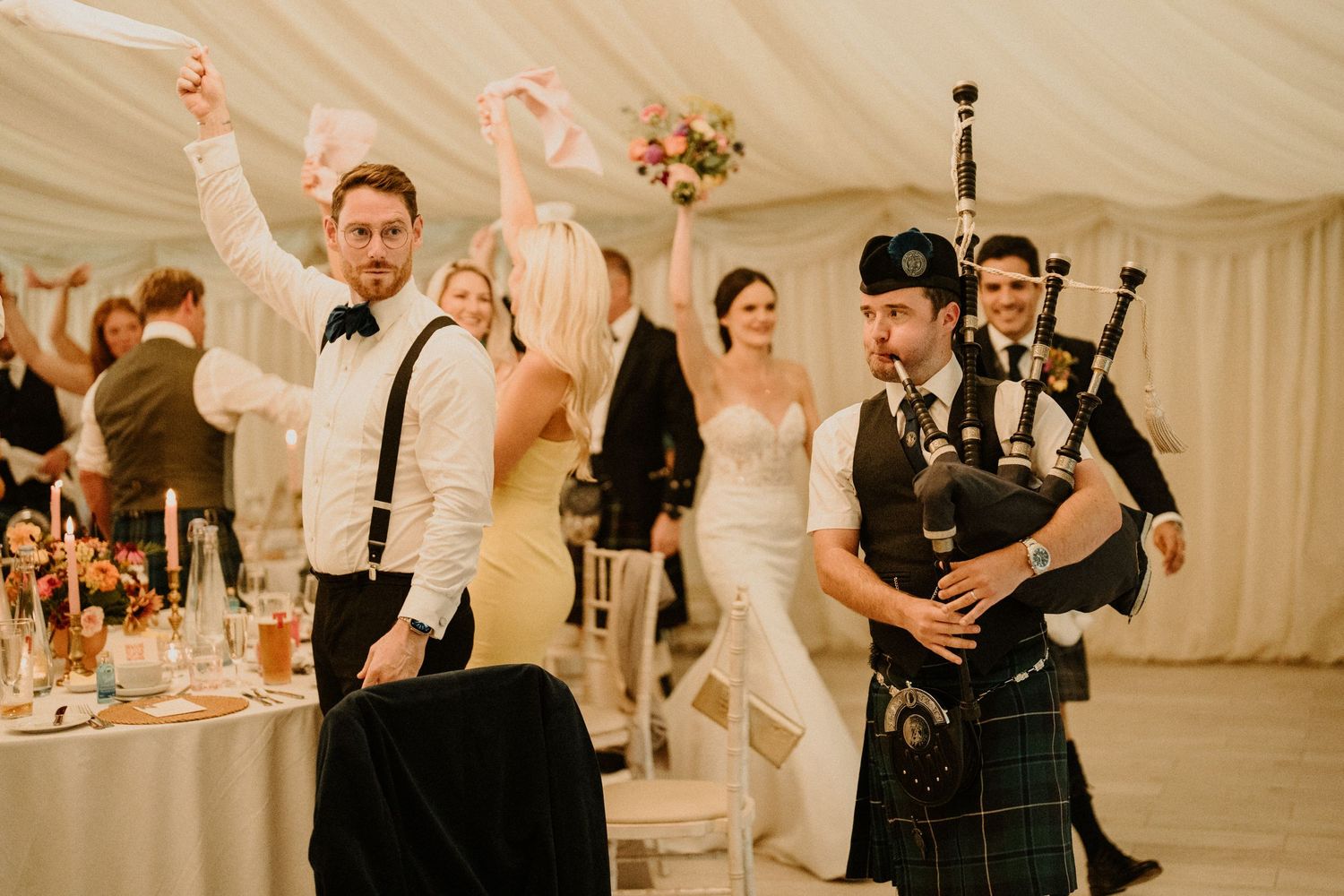 Wedding reception guests celebrate in a white draped tent decorated with floral arrangements and warm lighting.