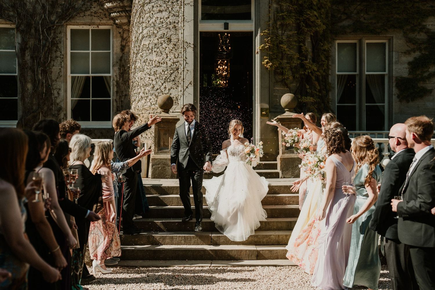 Wedding celebration outside a historic stone building with guests throwing confetti.