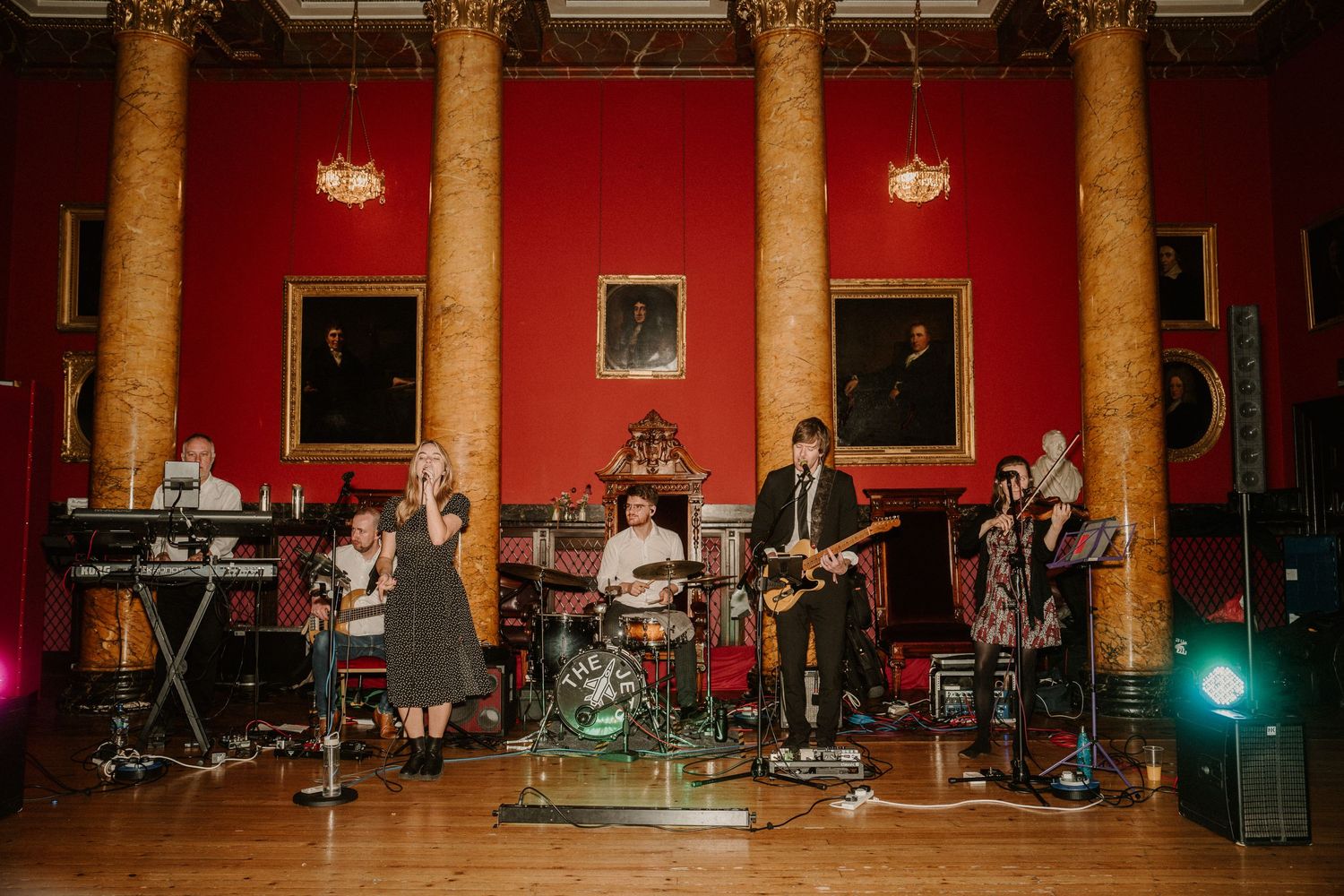Live band performs on stage in a red-walled venue with columns and lighting equipment.