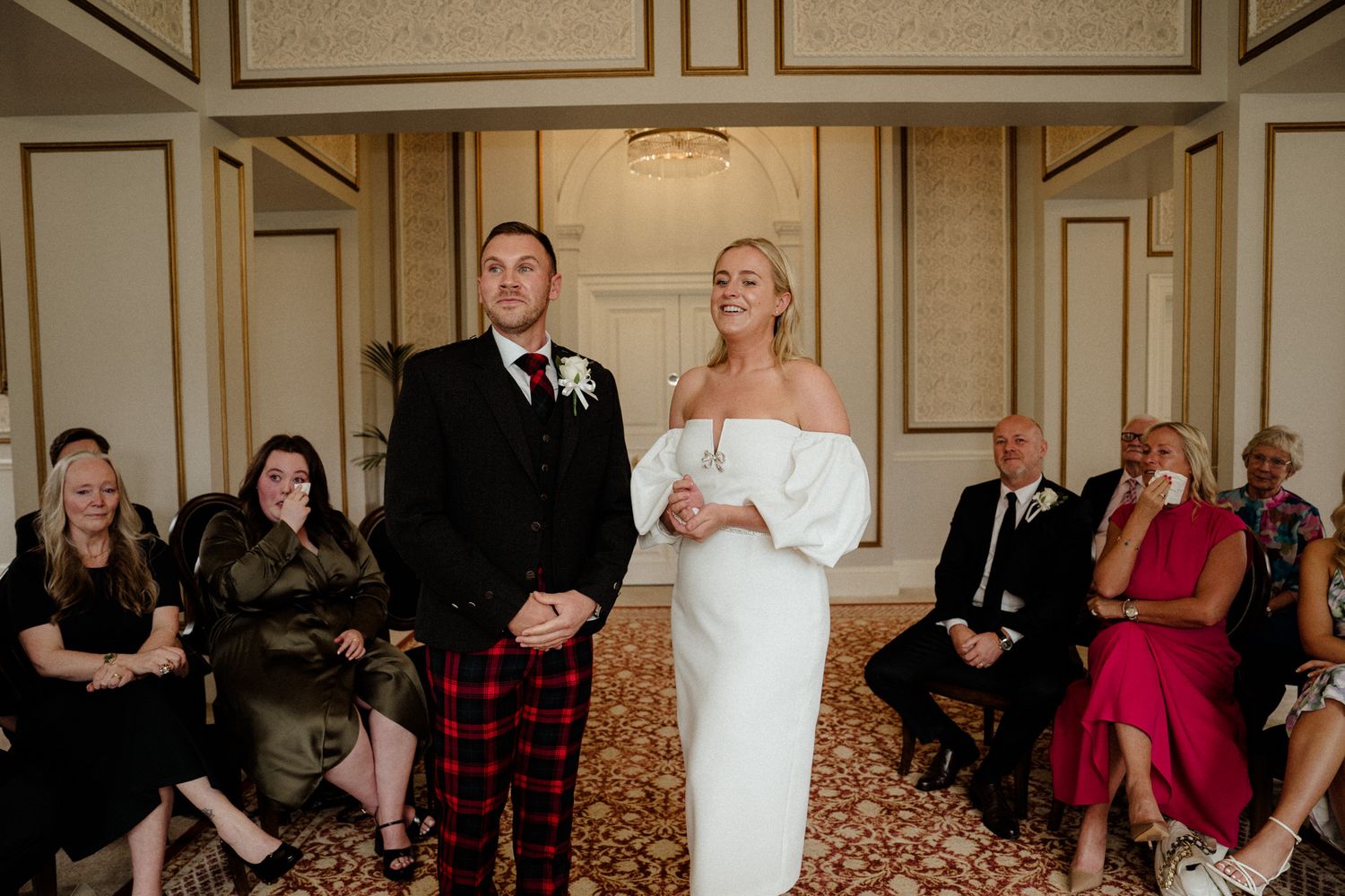 Wedding ceremony in a grand ballroom with ornate moldings and chandeliers.