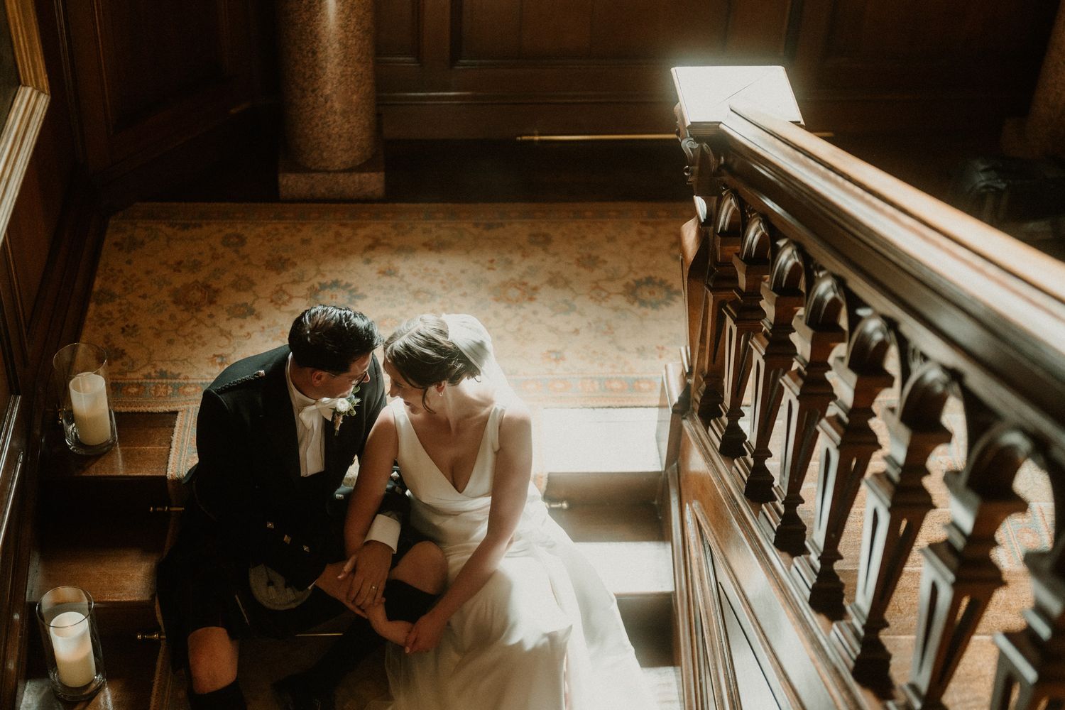 Bride in a white gown and groom in a kilt sit close together on a wooden staircase in a historic venue, bathed in warm natural light.
