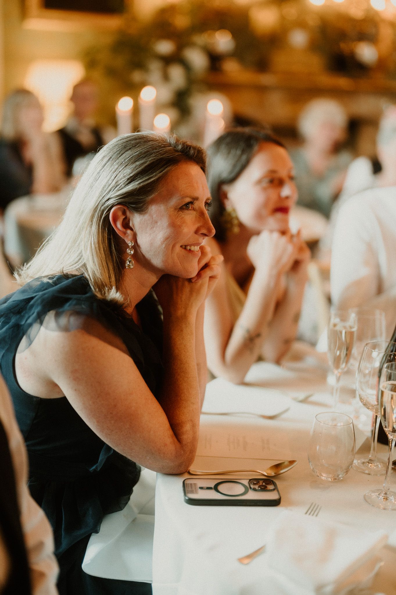 Smiling woman in a navy dress rests her chin on her hands at a candlelit wedding reception table while other guests look on.