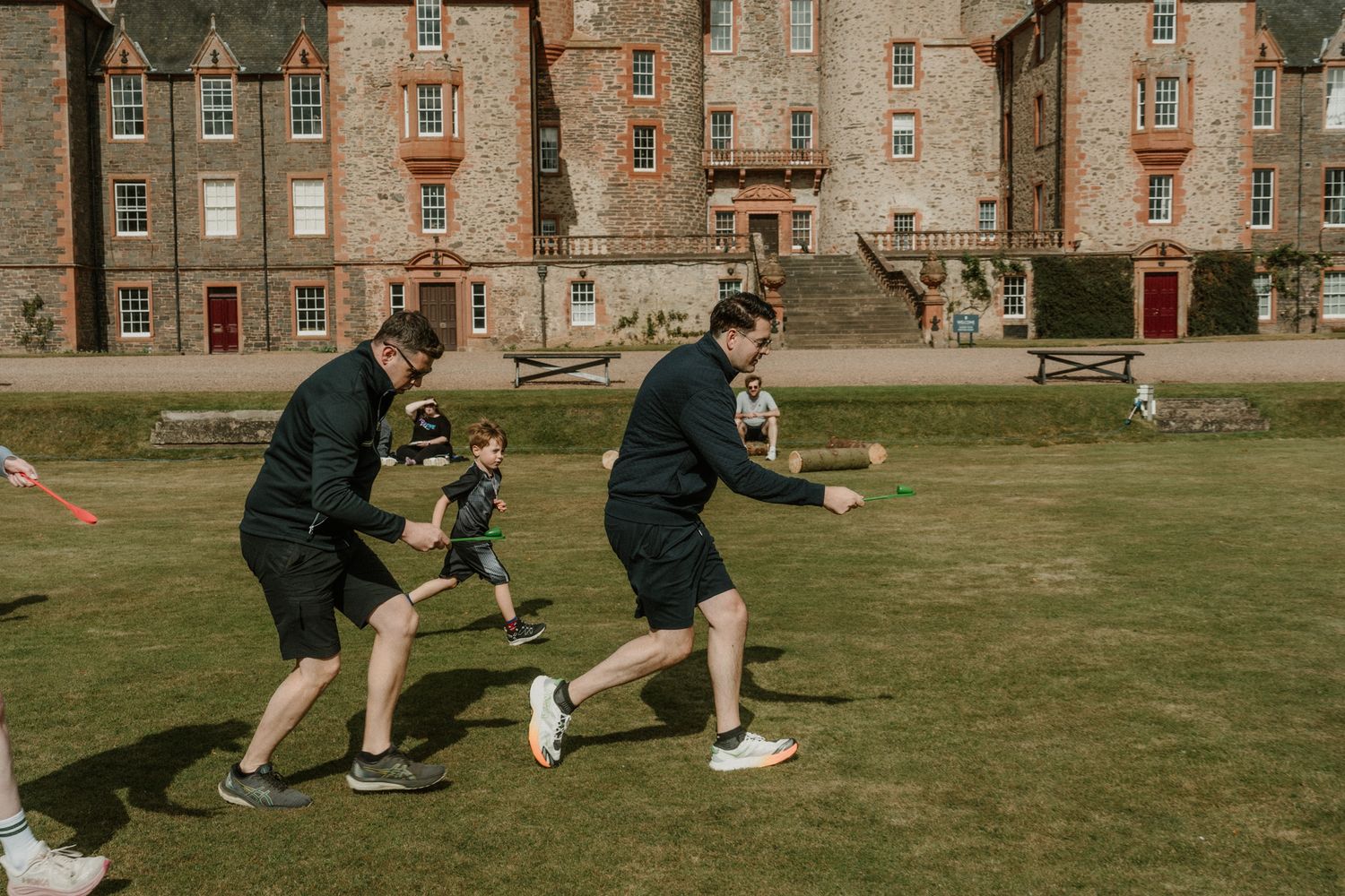 Two men in black athletic wear and a child play a game on a sunny lawn in front of a historic stone castle with turrets.