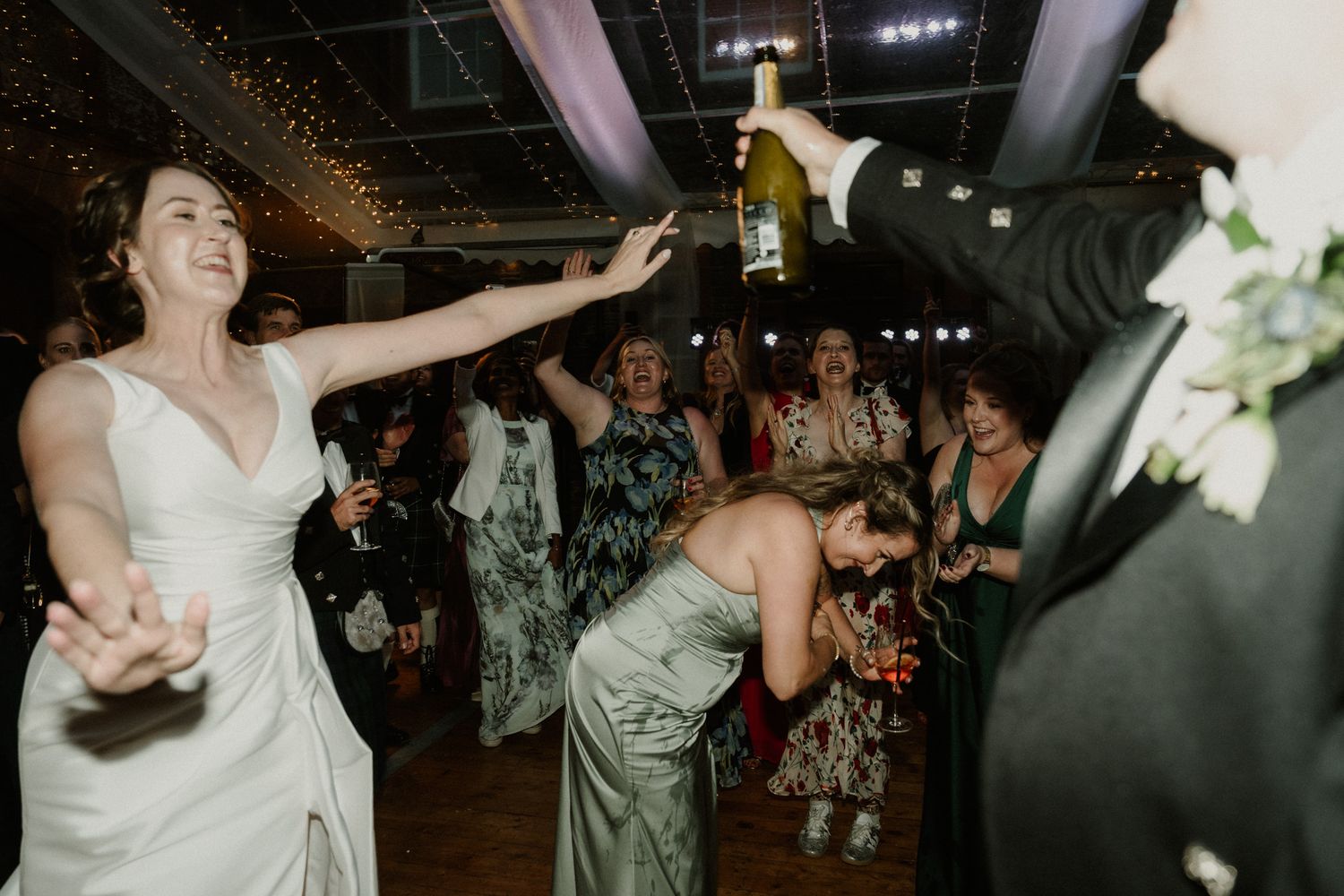 Bride in a white gown dances joyfully with wedding guests as a groom in a tuxedo holds up a champagne bottle at a lively reception.