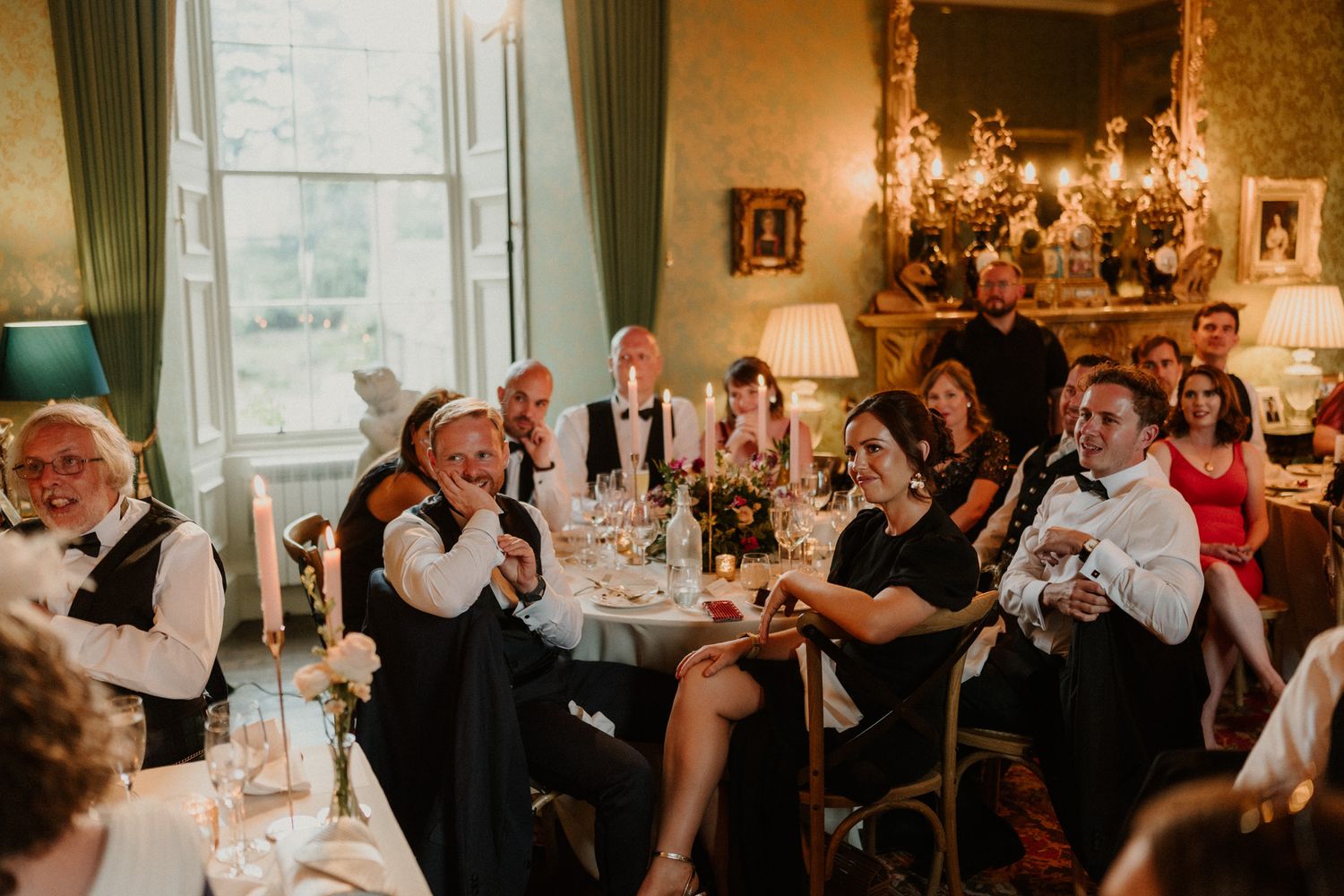 Smiling wedding guests in formal attire laugh and applaud around candlelit dining tables in an elegant ballroom with ornate decor.