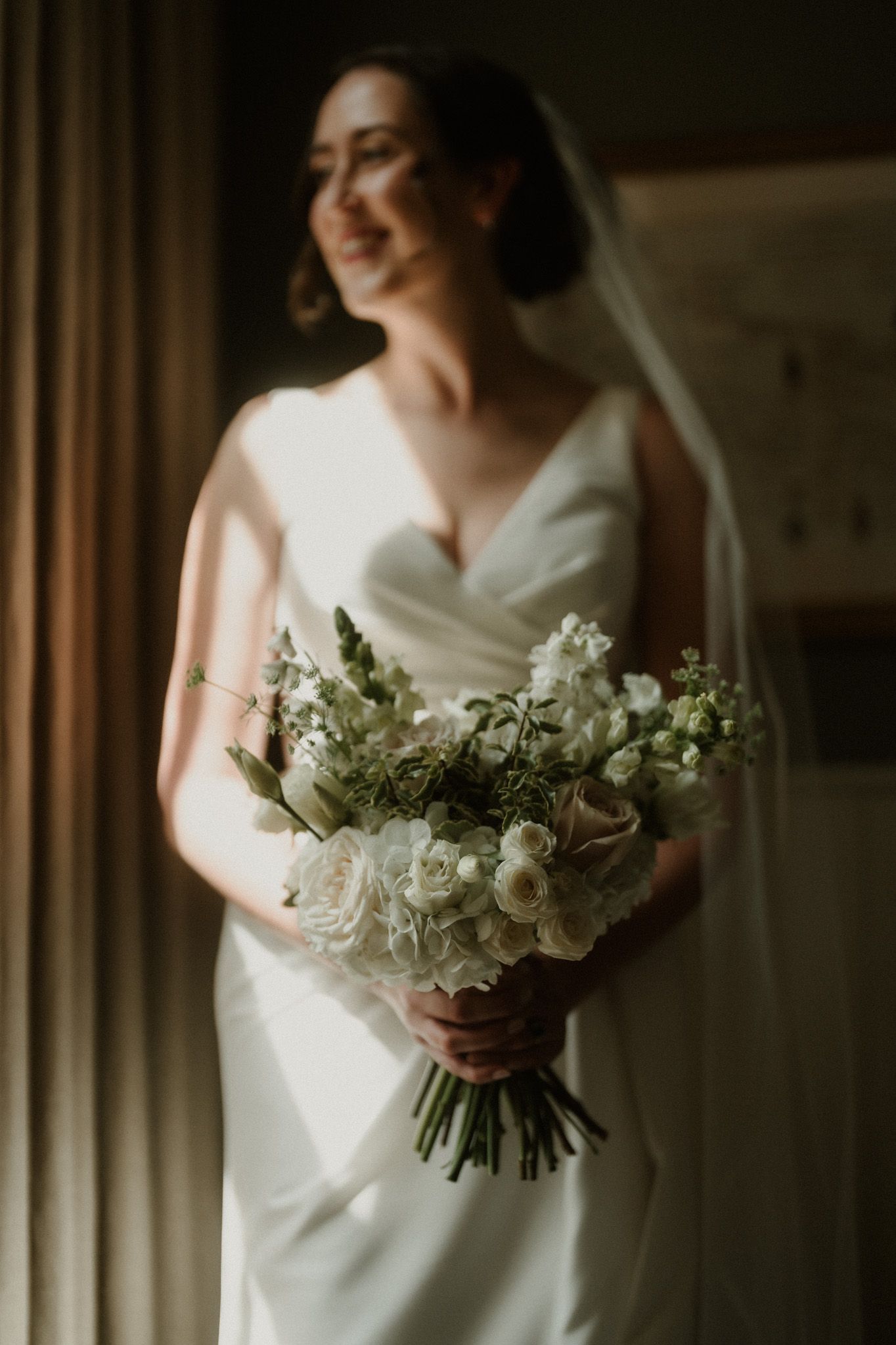 A smiling bride in an elegant white V-neck gown and veil holds a white rose and greenery bouquet, softly lit by window light.