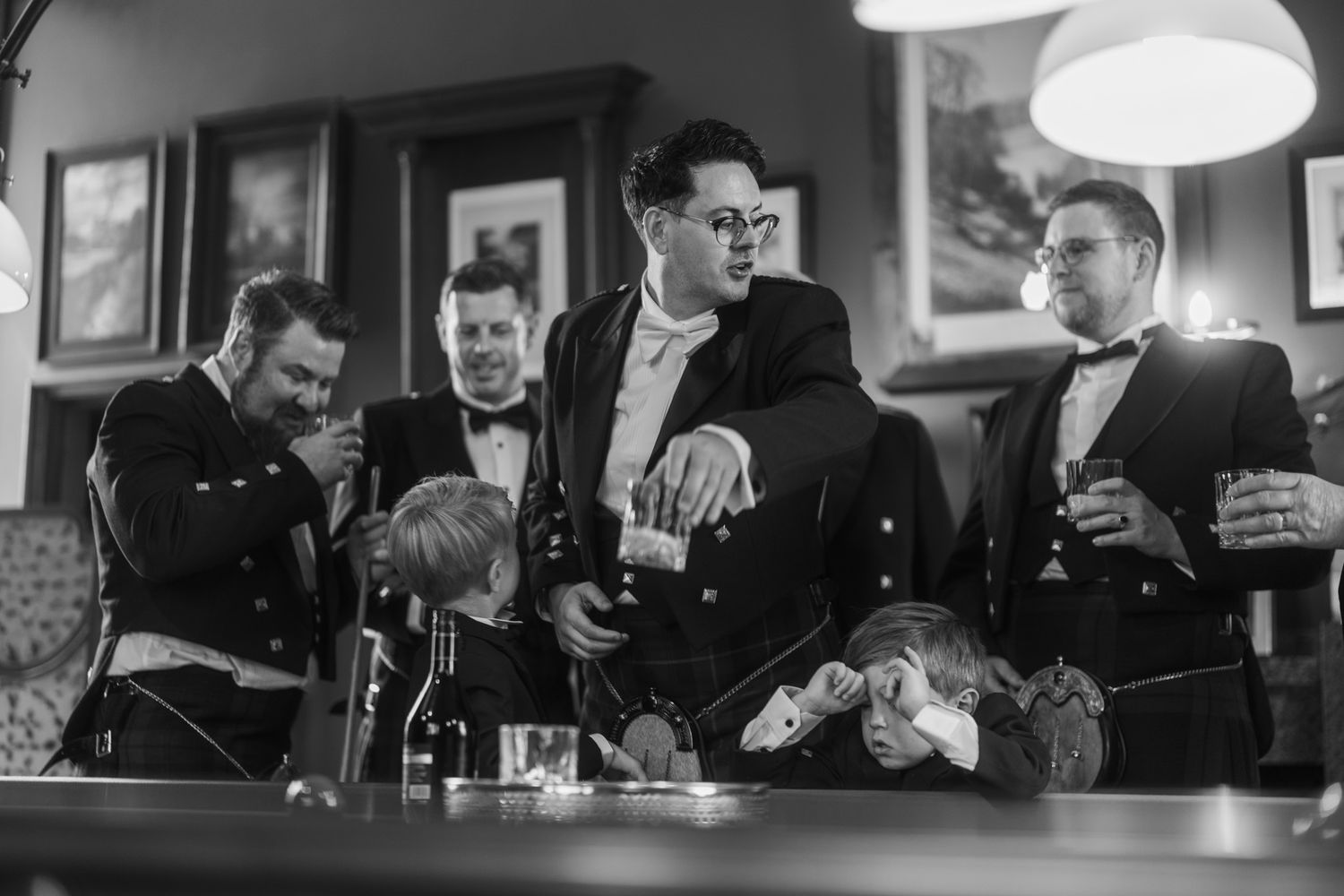 Groomsmen in black formal wear and bow ties laugh and share drinks around a wooden bar before the wedding ceremony.