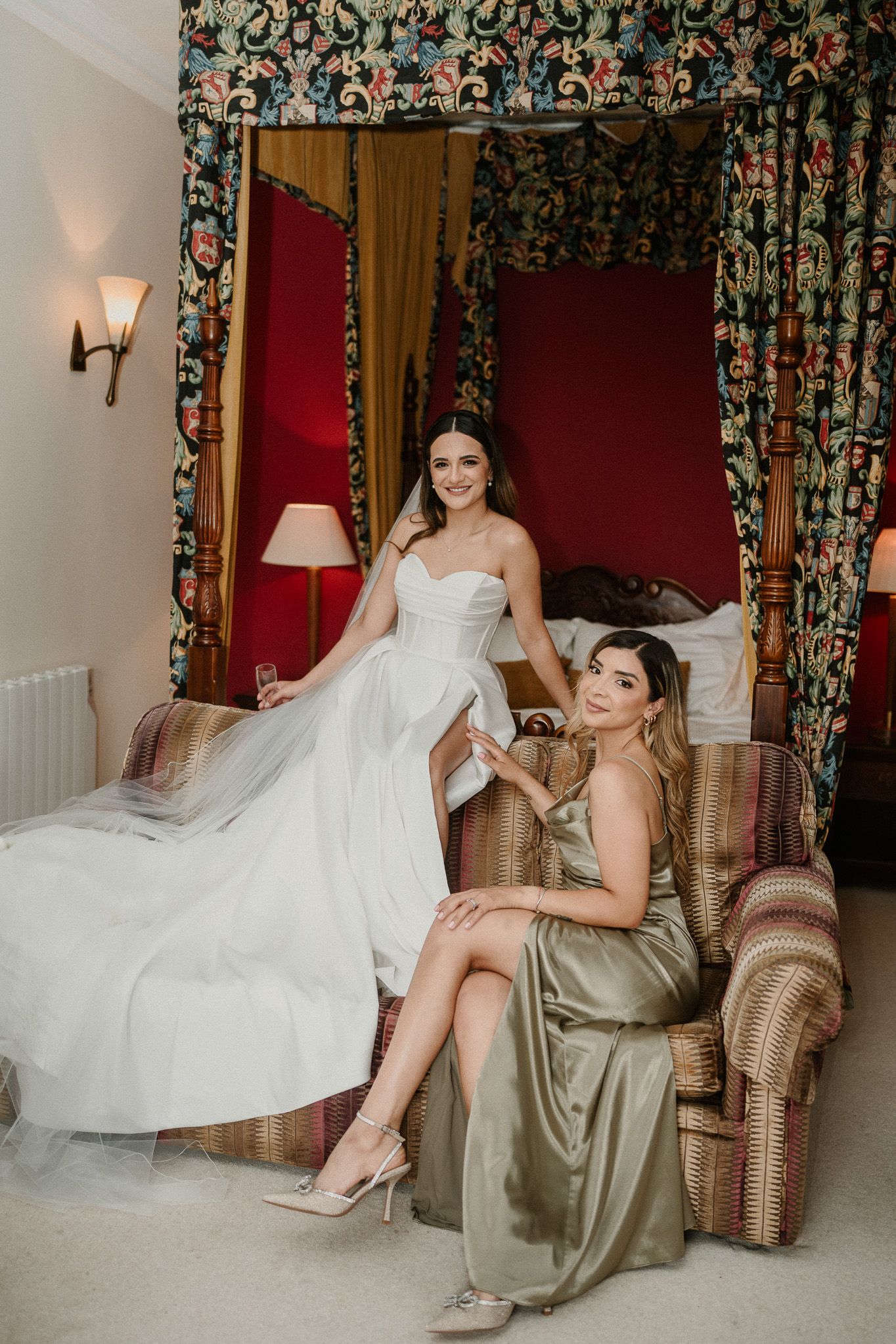 Bride in white gown and bridesmaid in sage satin dress pose elegantly in a luxurious four-poster bed room.