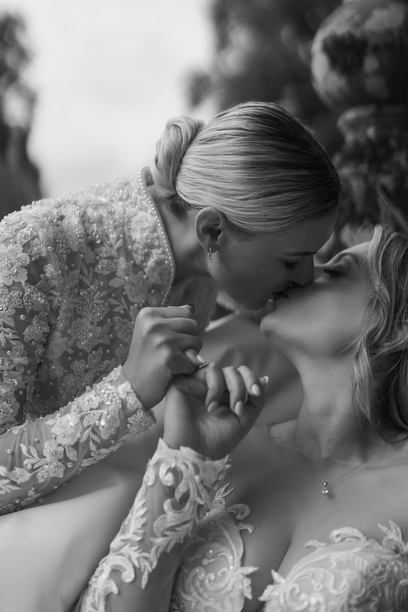 Two brides in lace wedding gowns share a romantic kiss while holding hands in a black and white wedding photo.