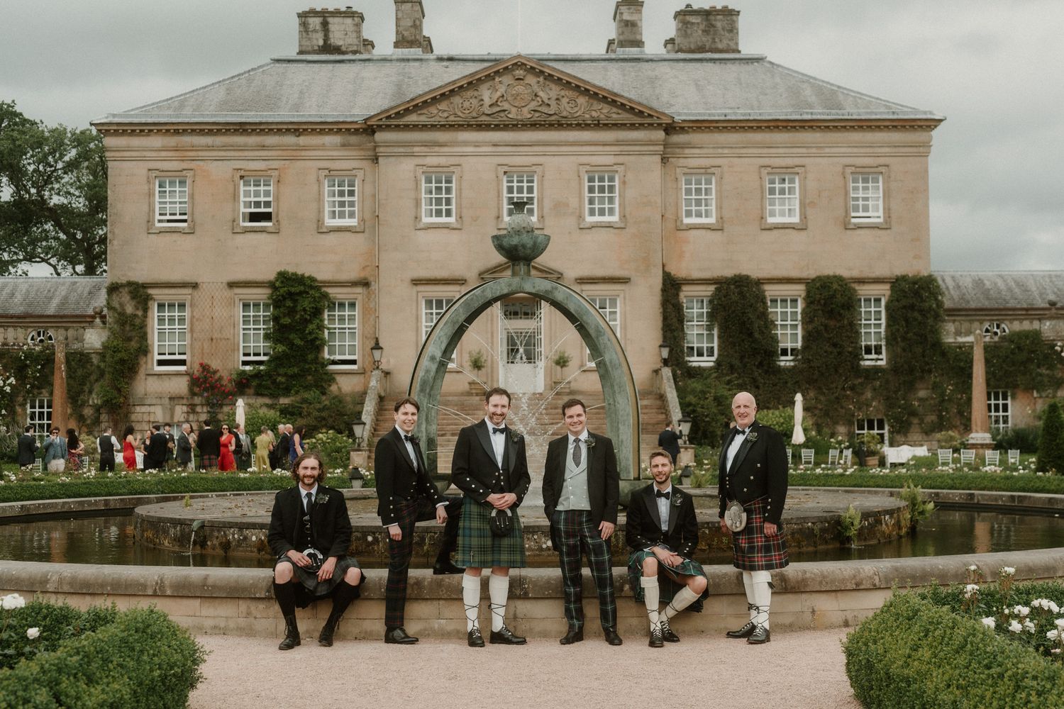 Groom and groomsmen in kilts posing by a fountain outside a grand Scottish estate wedding venue.