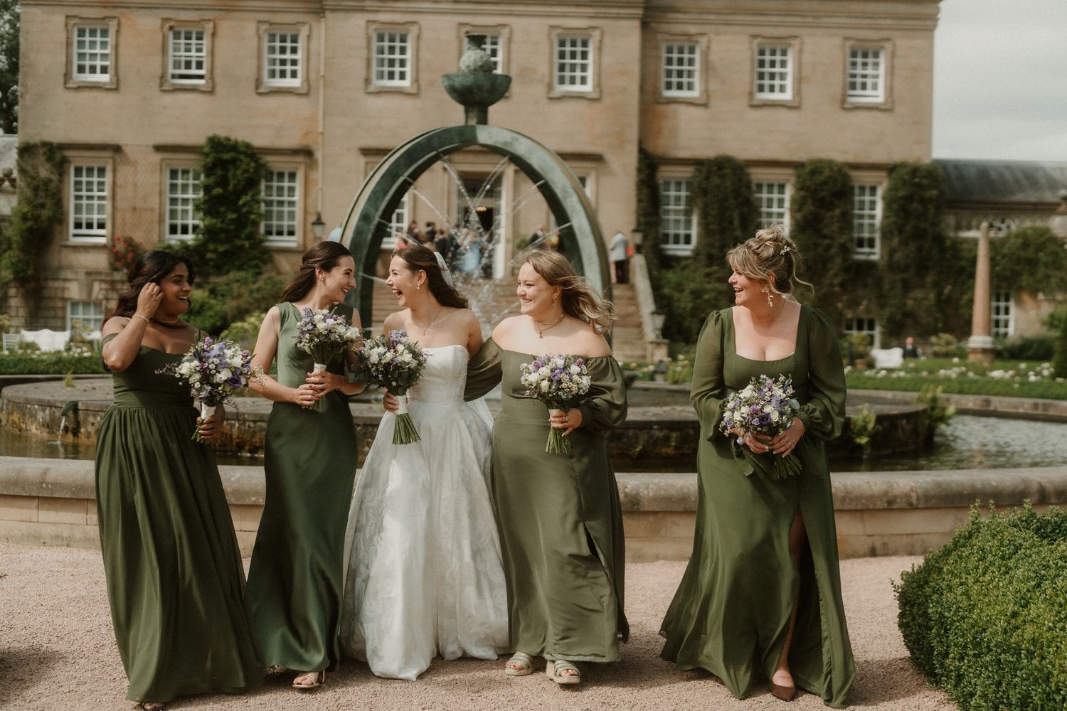 Bride in white gown walks with four bridesmaids in olive green dresses, holding bouquets outside an elegant manor.