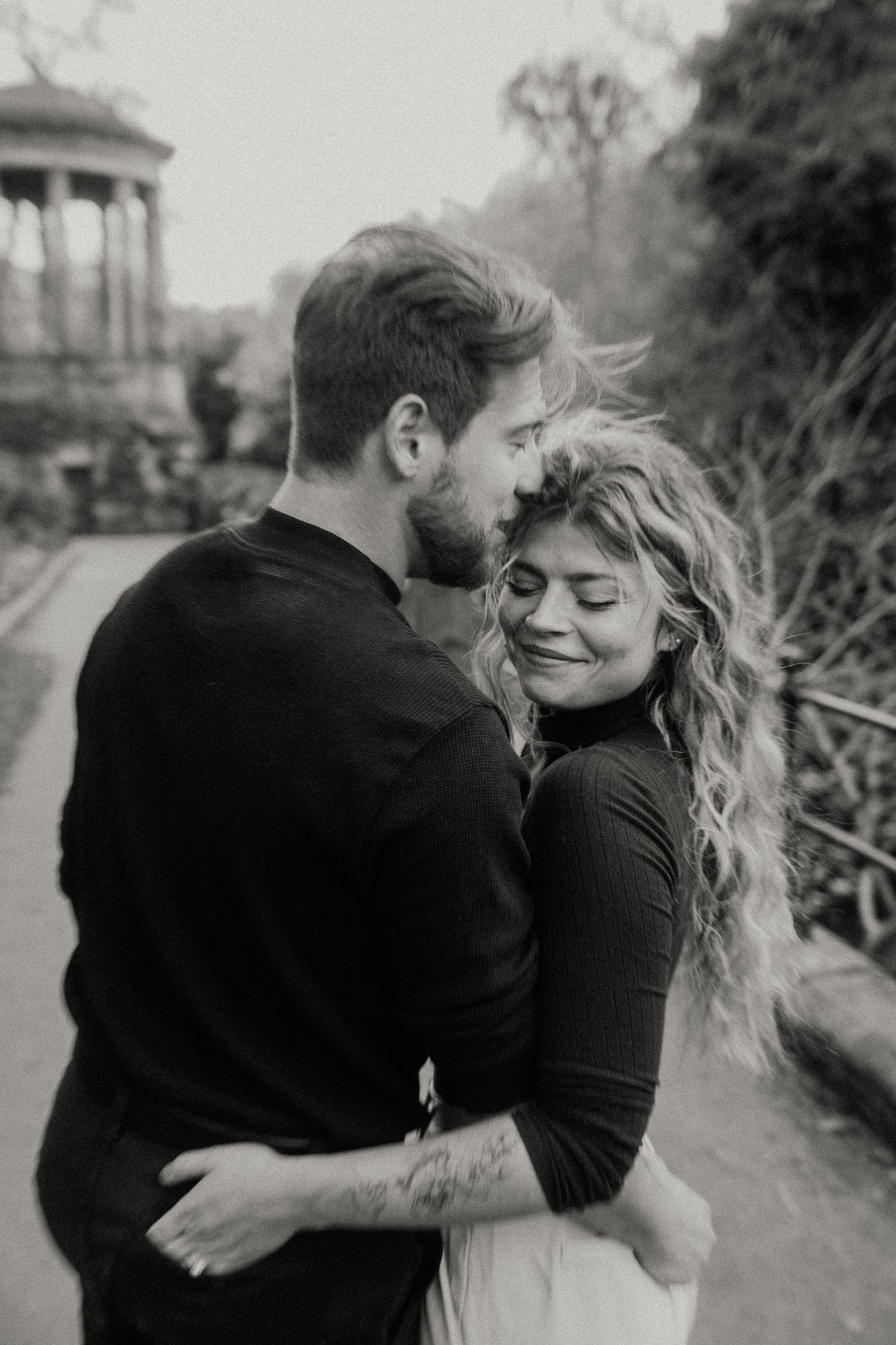 Black and white photo of a couple embracing tenderly in a park, woman smiling with eyes closed.