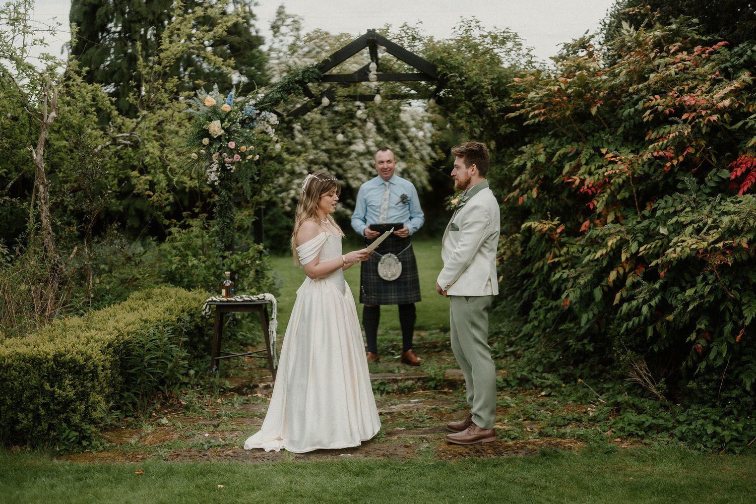 Couple exchanges vows outdoors under a rustic wooden arch surrounded by lush greenery with an officiant in a kilt.