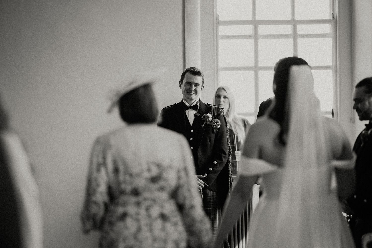 Wedding ceremony guests seated in rows at an elegant indoor venue with large windows and chandeliers.
