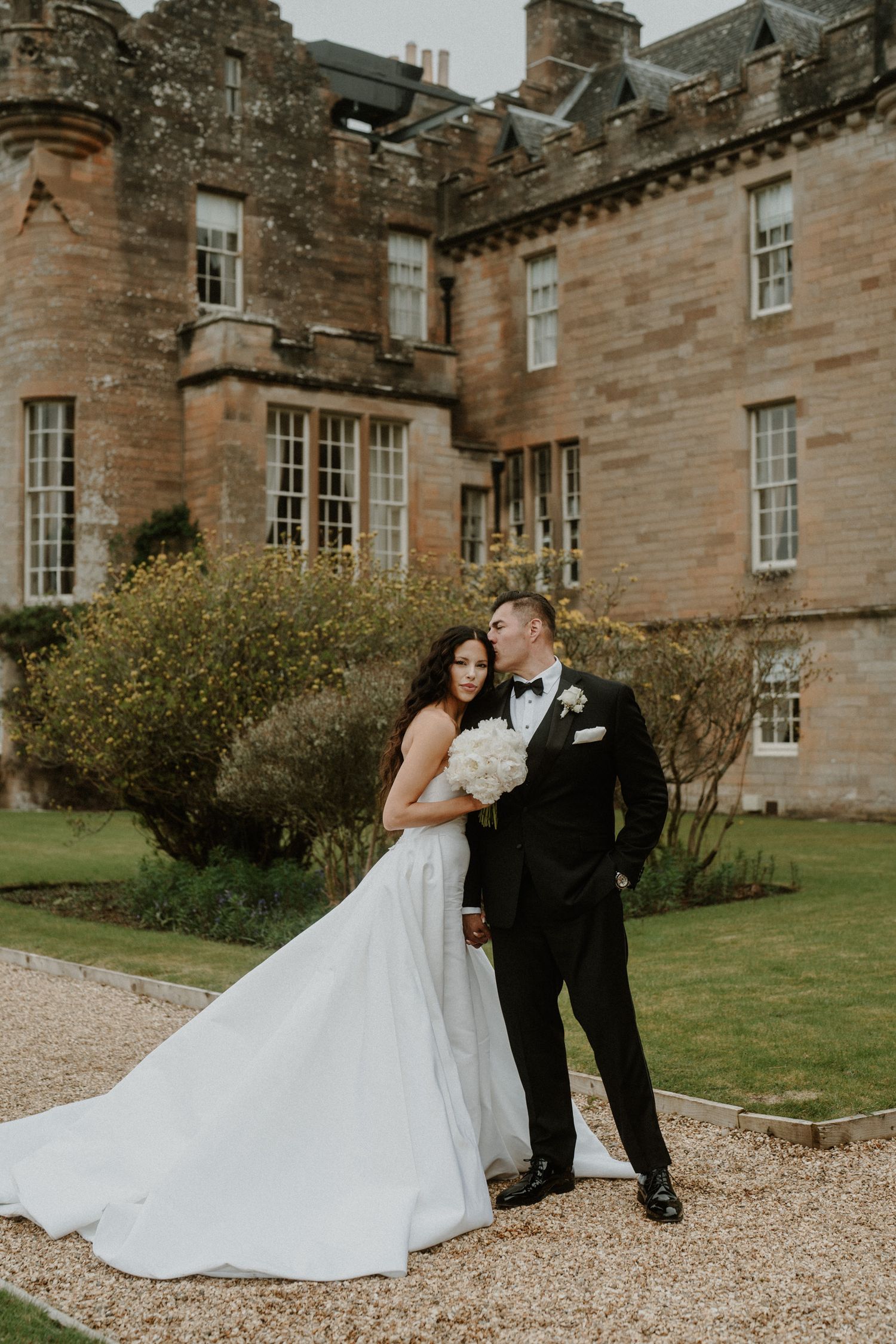 A wedding couple poses on the gravel driveway of a majestic Scottish castle.