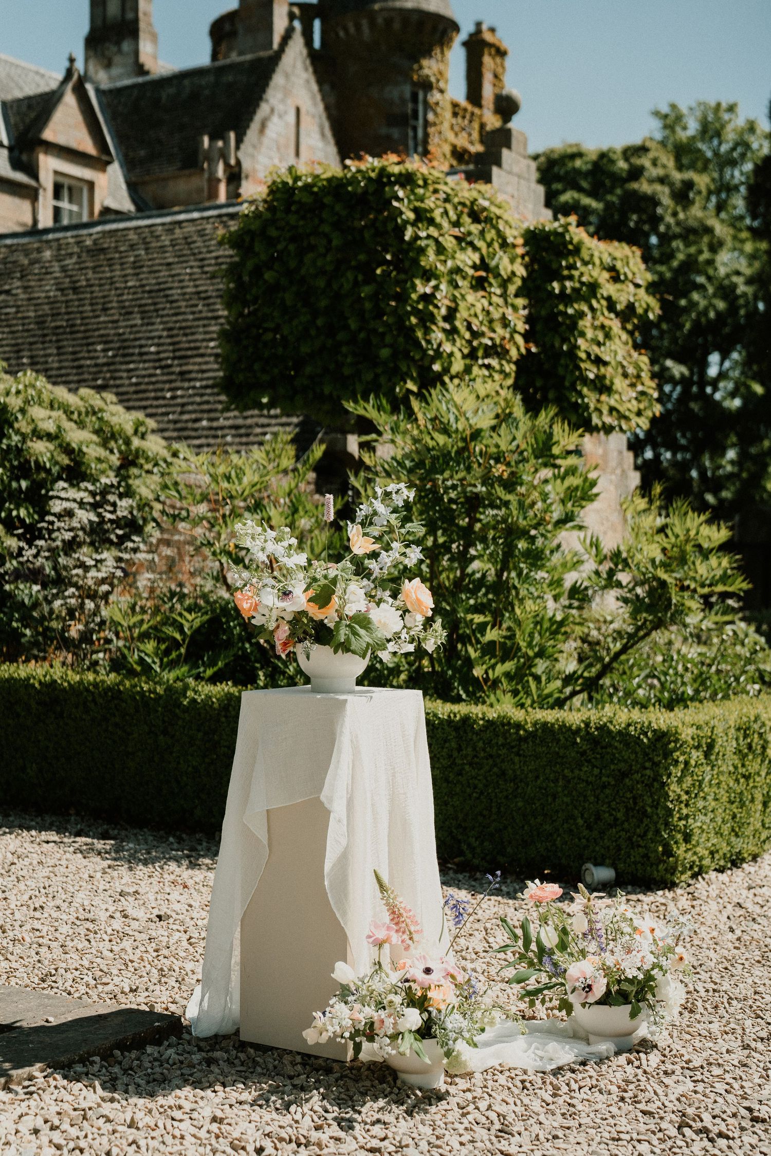 White floral arrangements in modern vases displayed on gravel path with garden backdrop.