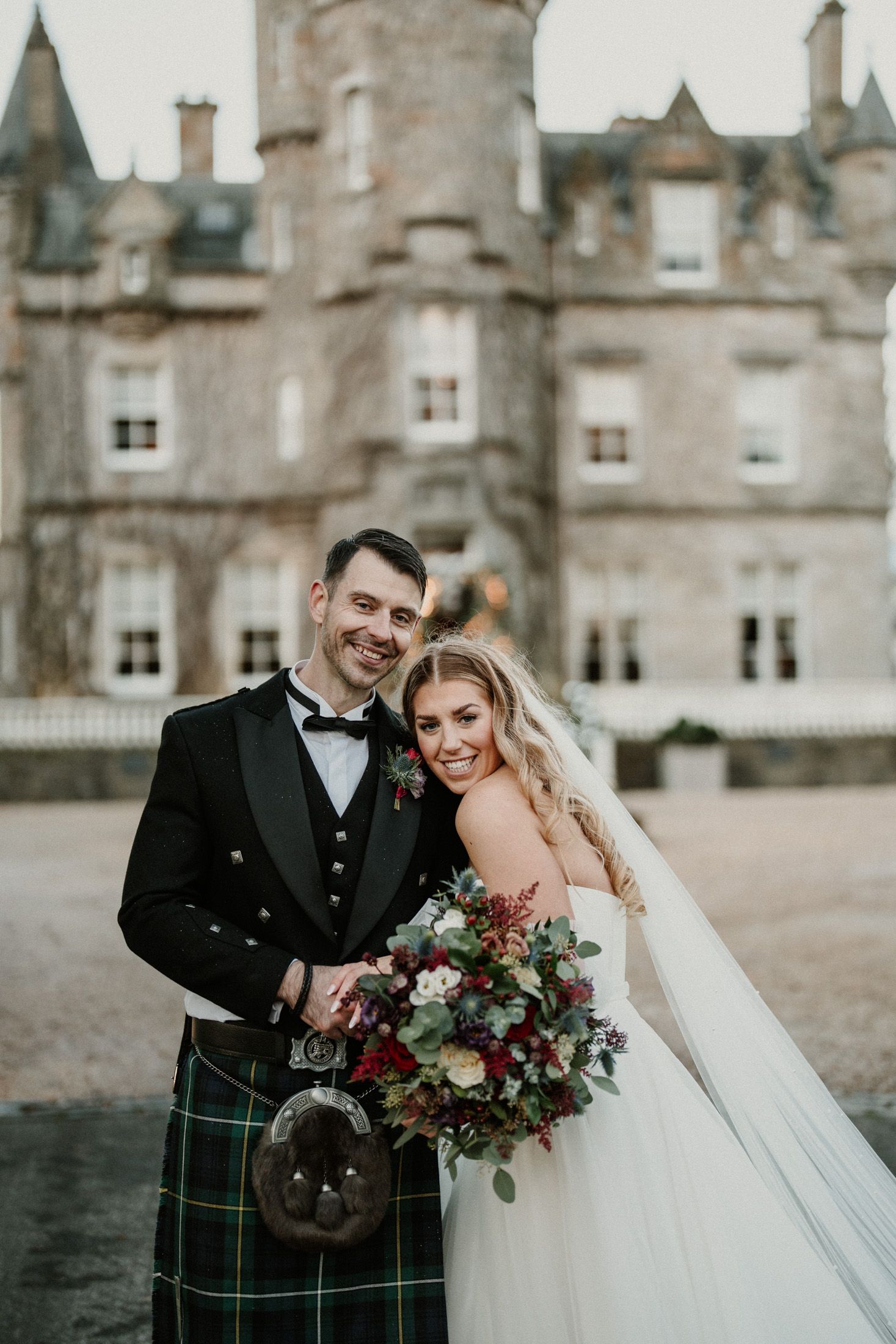 A romantic wedding photo shoot in front of a Scottish castle with the couple wearing traditional highland attire.