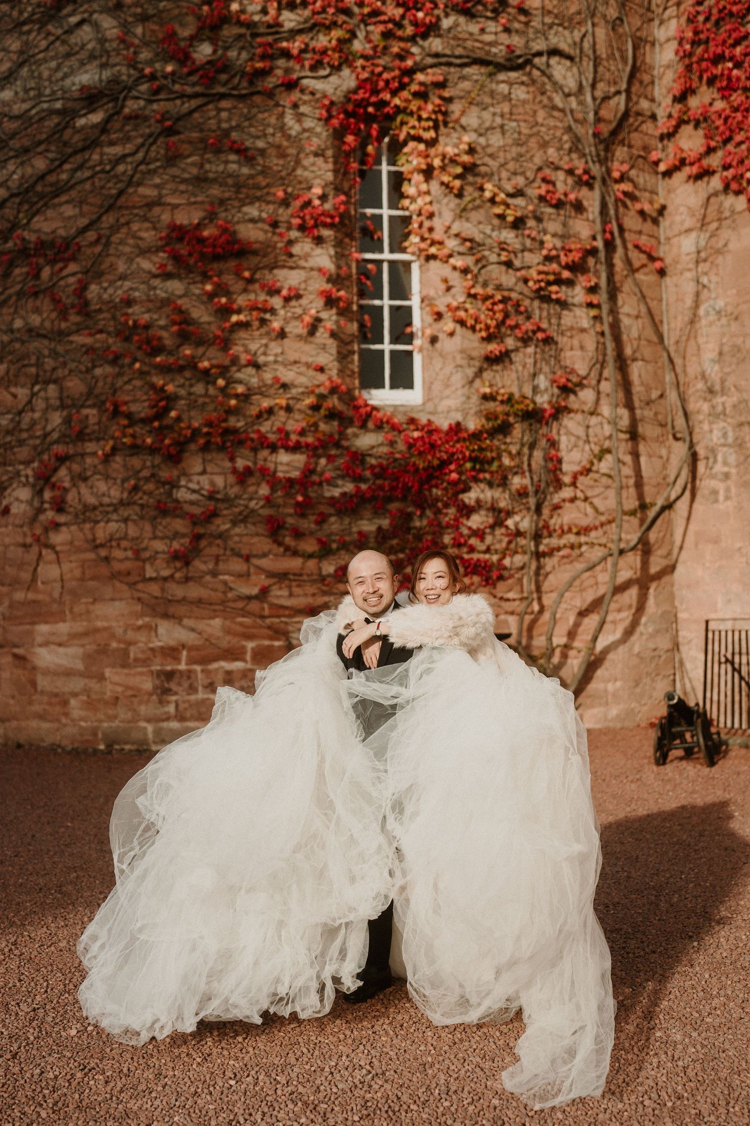 Two white wedding dresses spread across gravel against a brick wall with red ivy.
