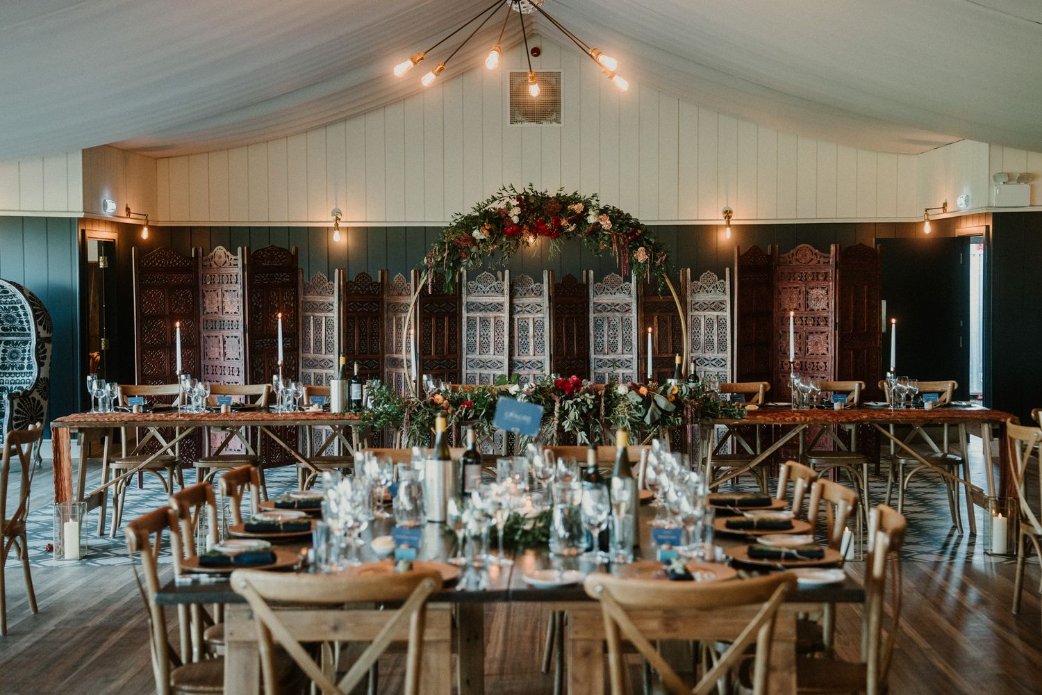 Elegant wedding reception venue with draped ceiling, wooden cross-back chairs, and long dining tables set for dinner.