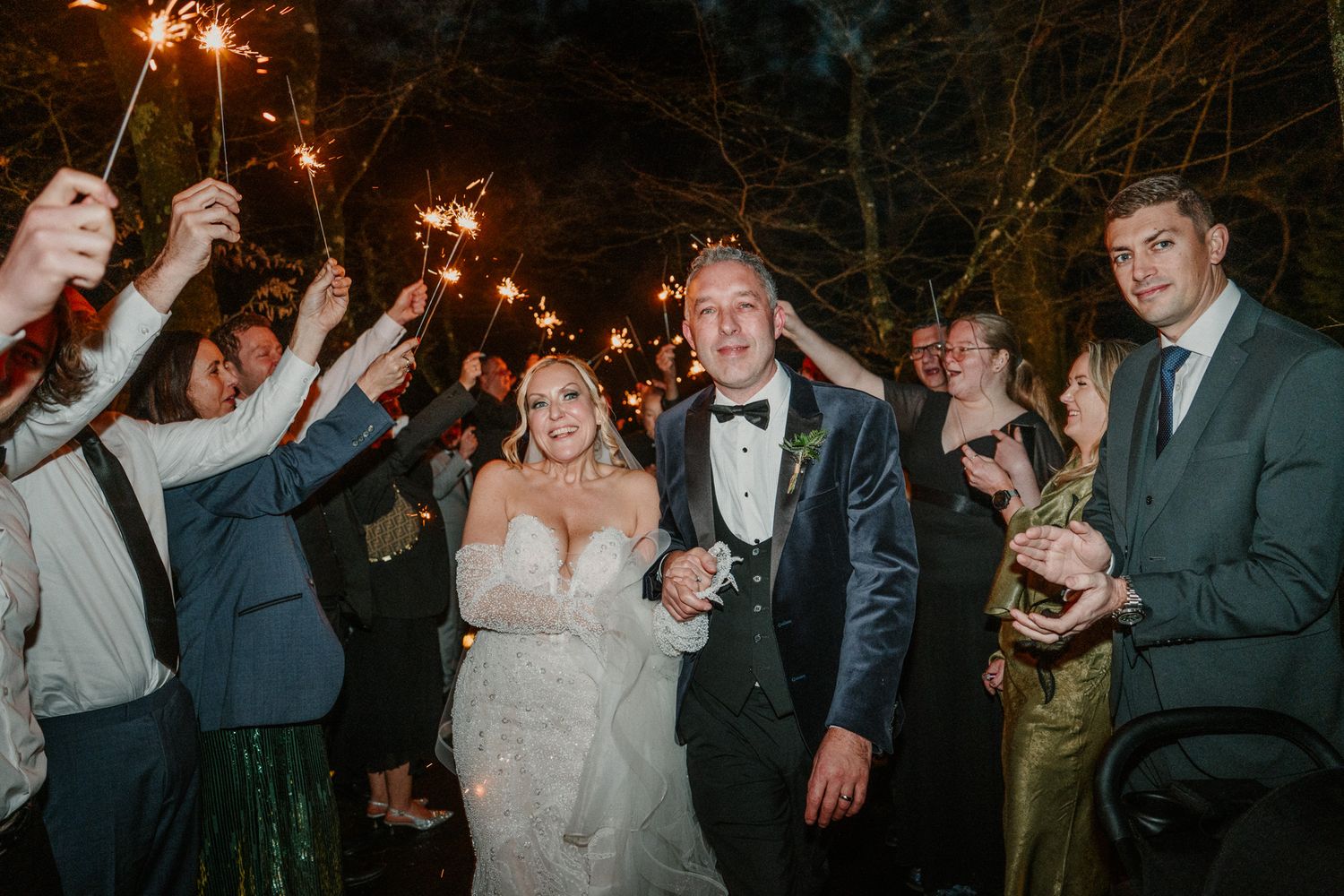 Wedding celebration with guests holding sparklers while dancing and cheering at night.