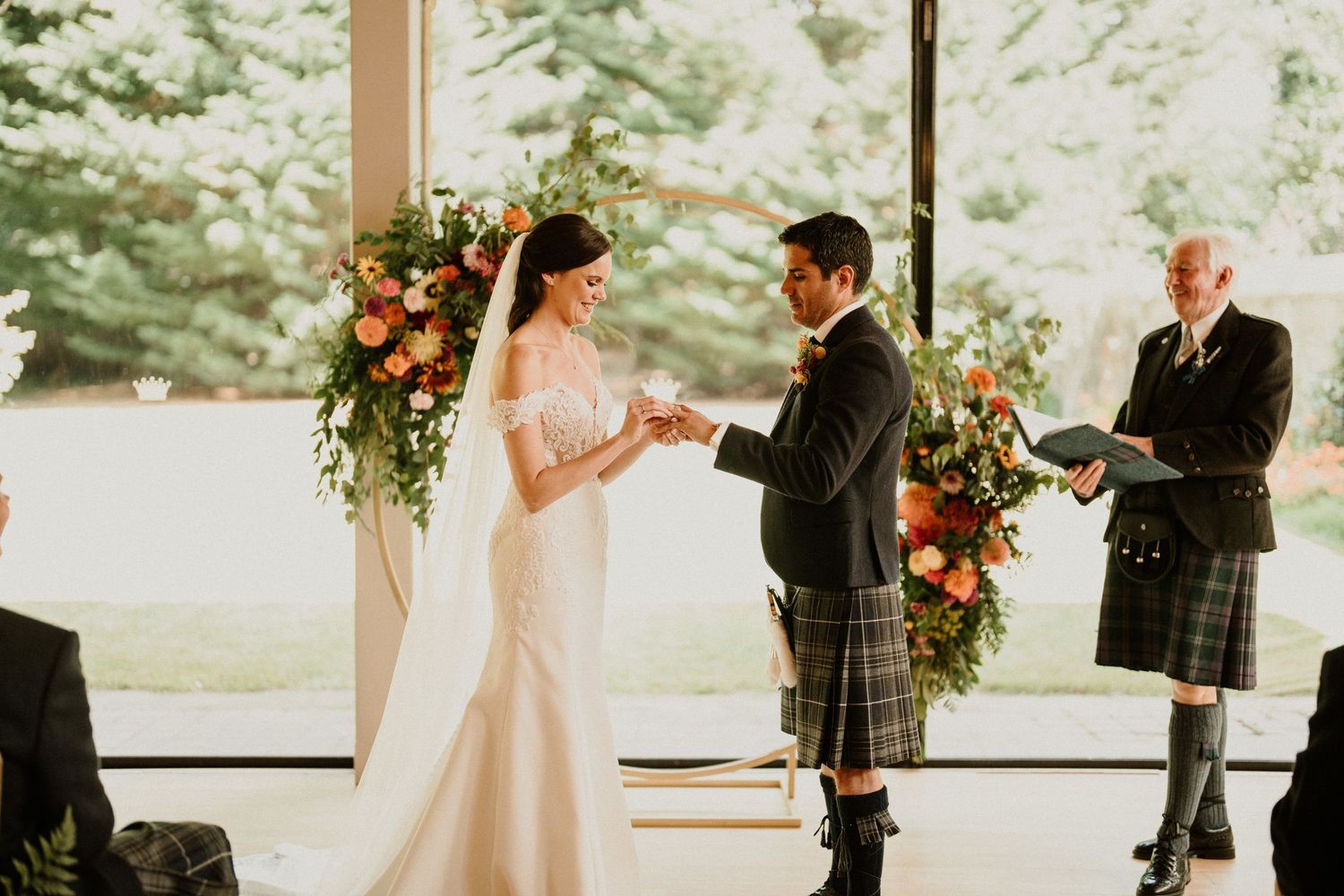 Wedding ceremony taking place in a bright indoor venue with floral decorations and natural lighting.