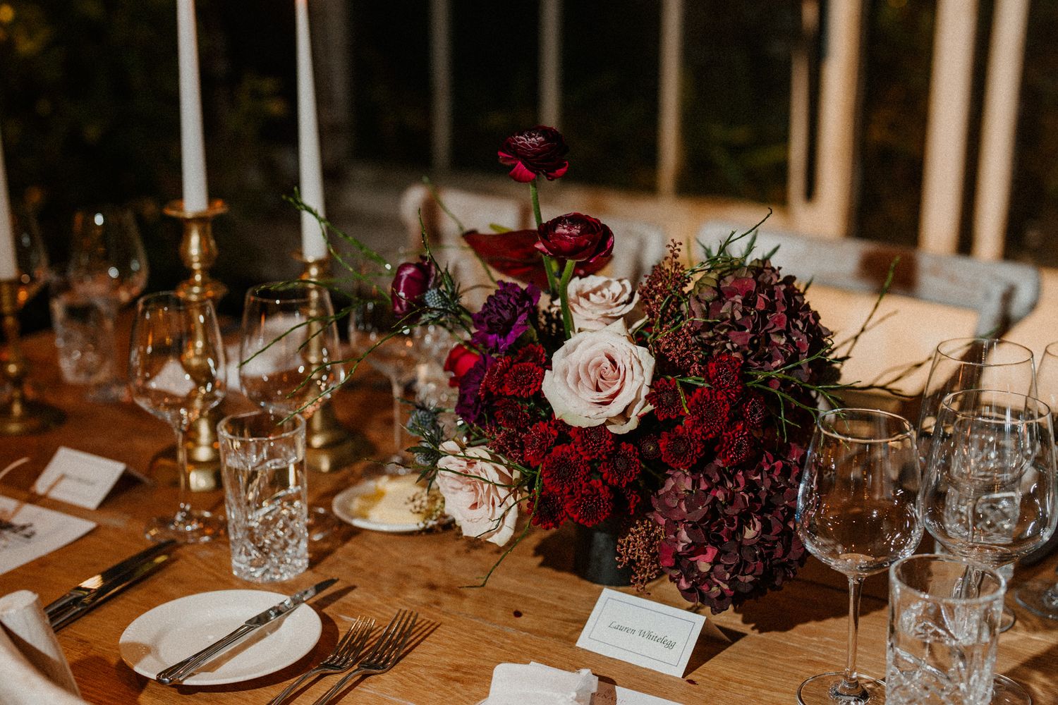 Wooden table decorated with burgundy and pink floral arrangements, crystal glasses, and elegant place settings.