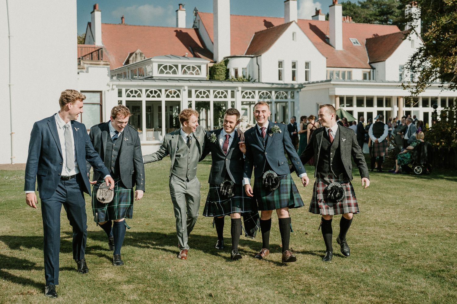 A group walks across a lawn wearing traditional Scottish formal wear and kilts.