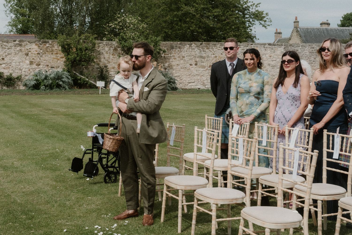 Man holding a baby stands beside wedding ceremony chairs outdoors while guests watch in a garden setting.