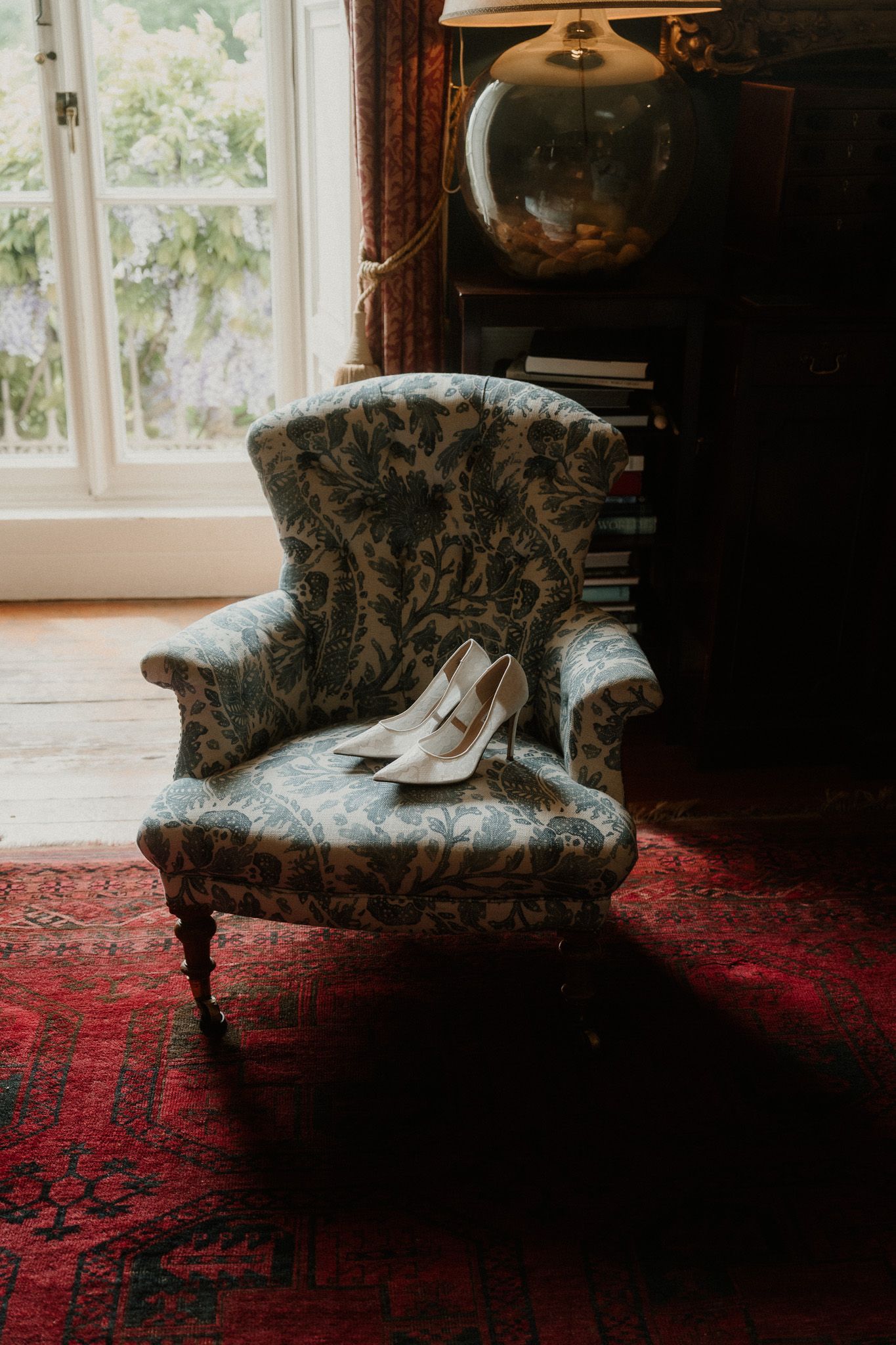 White bridal heels displayed on a vintage floral armchair atop a red Persian rug in an elegant, book-filled room.