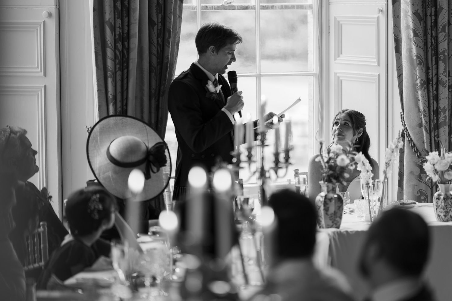 Black and white wedding reception photo of groom giving speech at elegant venue with guests seated at decorated tables.