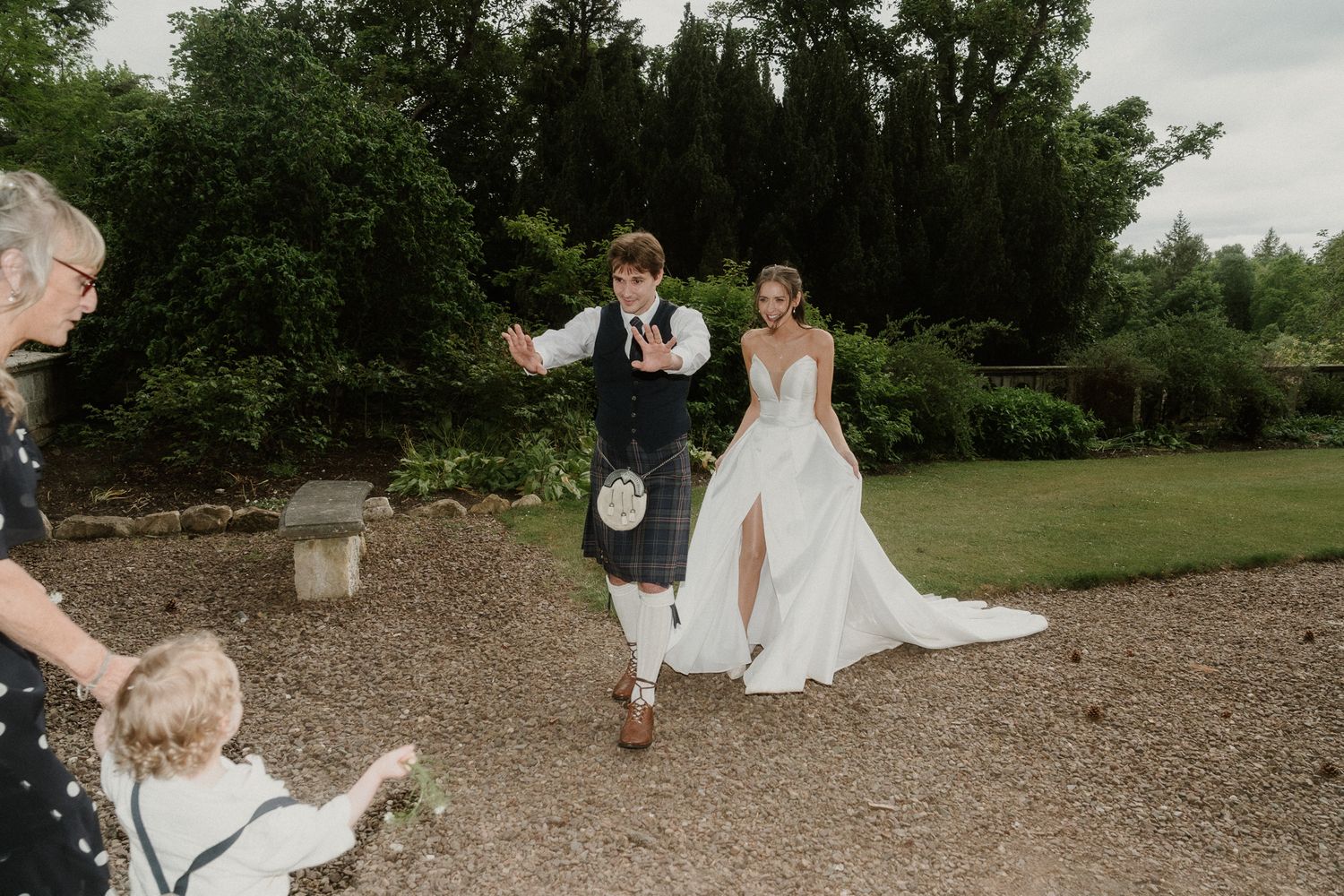 Bride in white gown and groom in Scottish kilt walk through garden as guests toss confetti at outdoor wedding celebration.