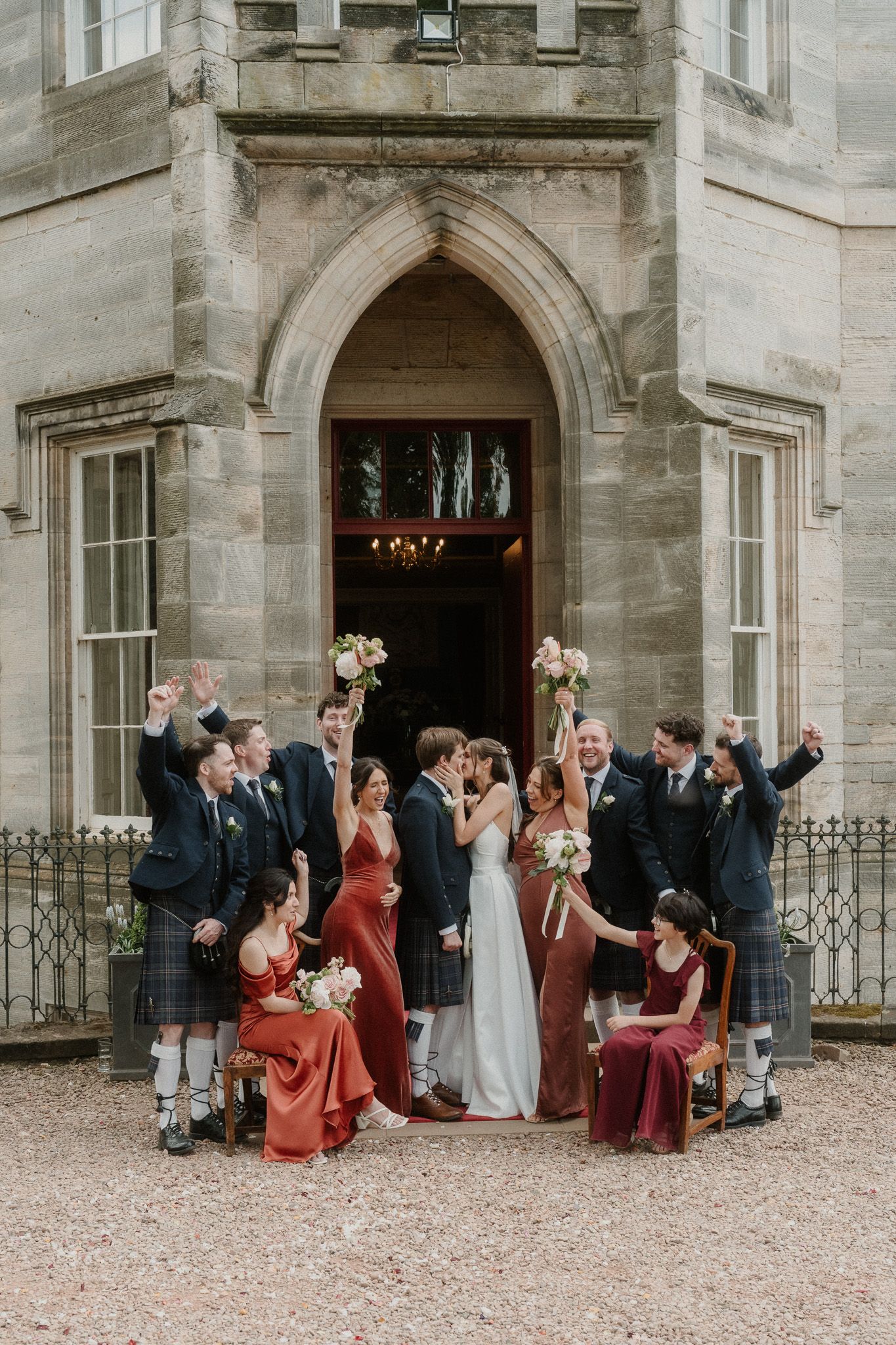 Joyful wedding party cheers outside a Scottish castle, with bridesmaids in red and groomsmen in traditional kilts.