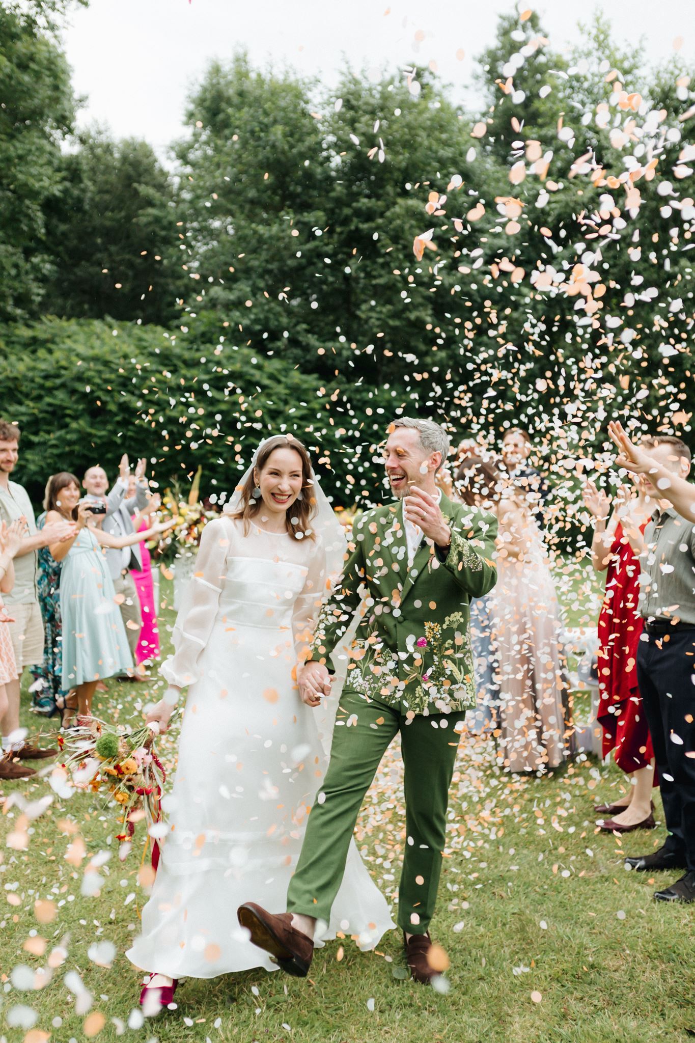 Bride and groom walk through confetti shower surrounded by cheering guests at outdoor wedding ceremony.