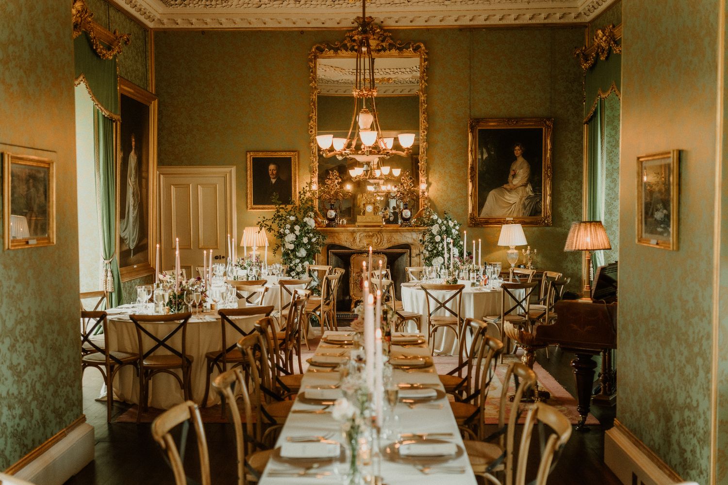 Elegant wedding dining room with long tables, candles, floral arrangements, gold chandelier, and green wallpaper in a stately manor.