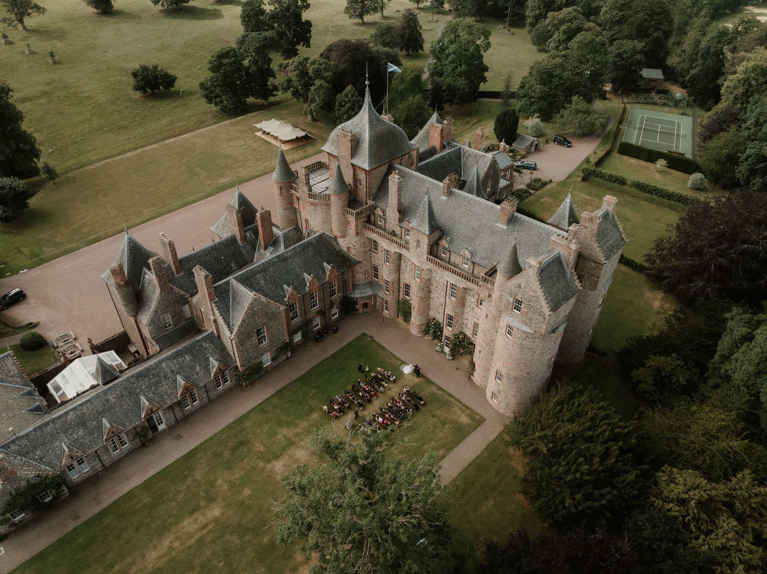 Aerial view of a historic Scottish baronial castle with turrets and slate roofs, surrounded by manicured lawns, gardens, and mature trees.