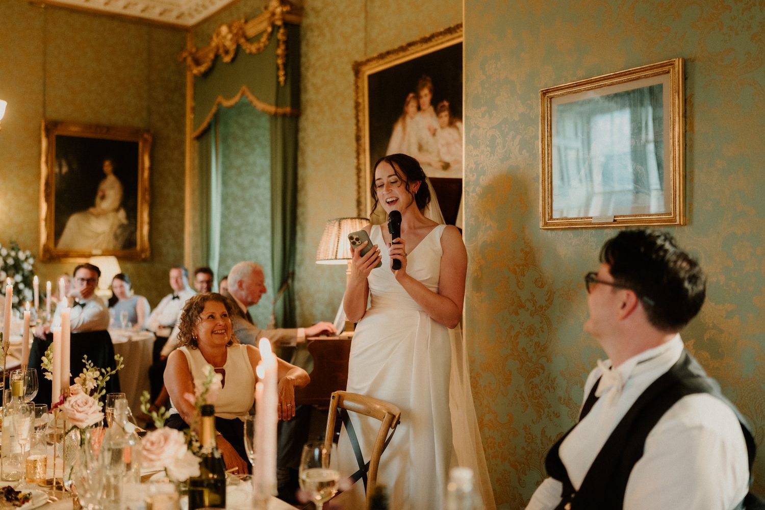 A bride in a white gown holds a microphone and gives a speech at an elegant wedding reception as seated guests listen.