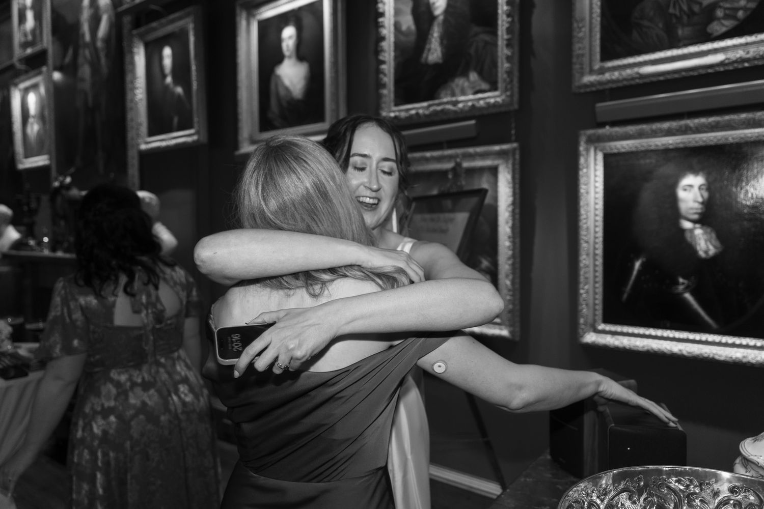 Two women share a warm hug in a gallery room lined with framed vintage portrait paintings in a black and white photo.