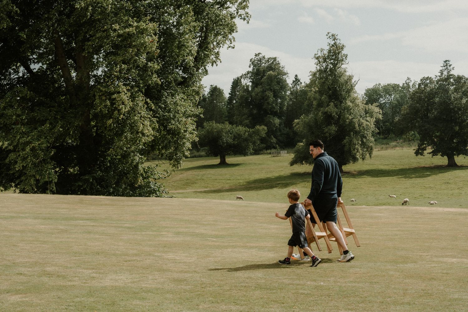 A man and a young child walk across a sunny, open grass field with tall green trees in the background.