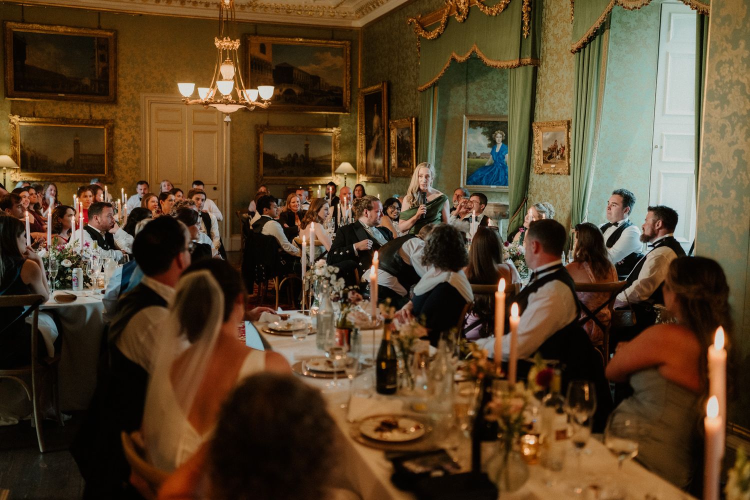 Wedding guests seated at long candlelit tables with glassware and flowers in an elegant ballroom with green walls and gilded framed art.