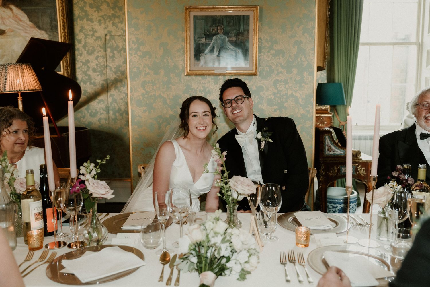 Smiling bride and groom sit at an elegant wedding reception table with candles, flowers and wine glasses in a classic room with framed art.
