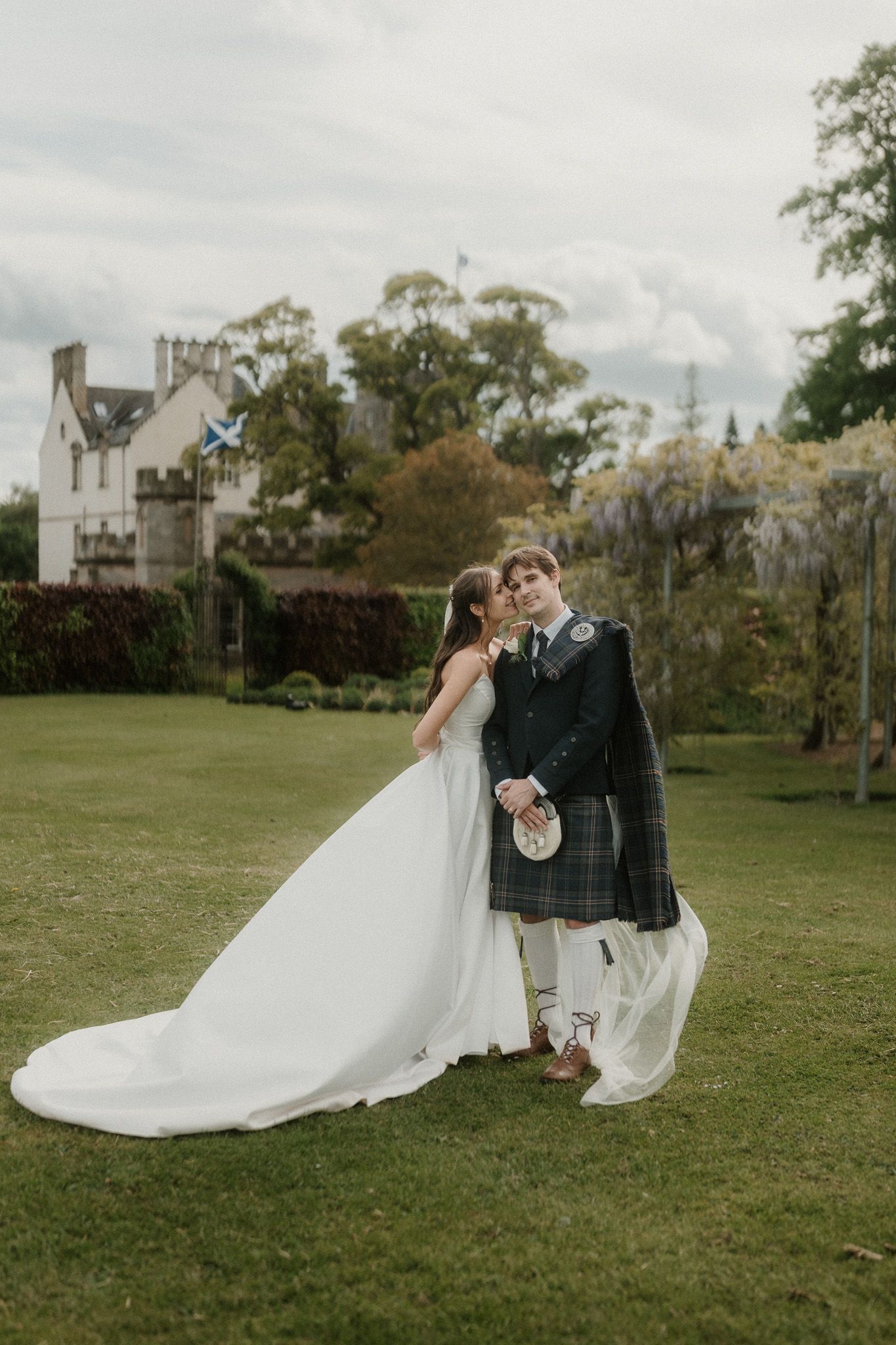 Bride in white gown and groom in Scottish tartan kilt embrace on green lawn with historic manor house backdrop.