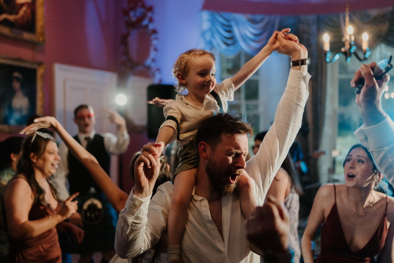 A joyful child sits on a man's shoulders at a lively wedding reception, surrounded by celebrating guests.