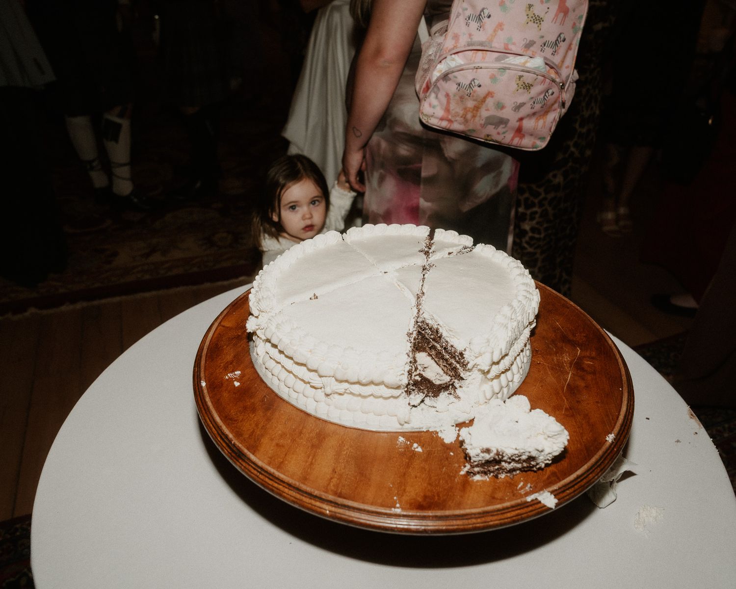 A young girl peeks behind a white frosted celebration cake on a wooden stand at a party.