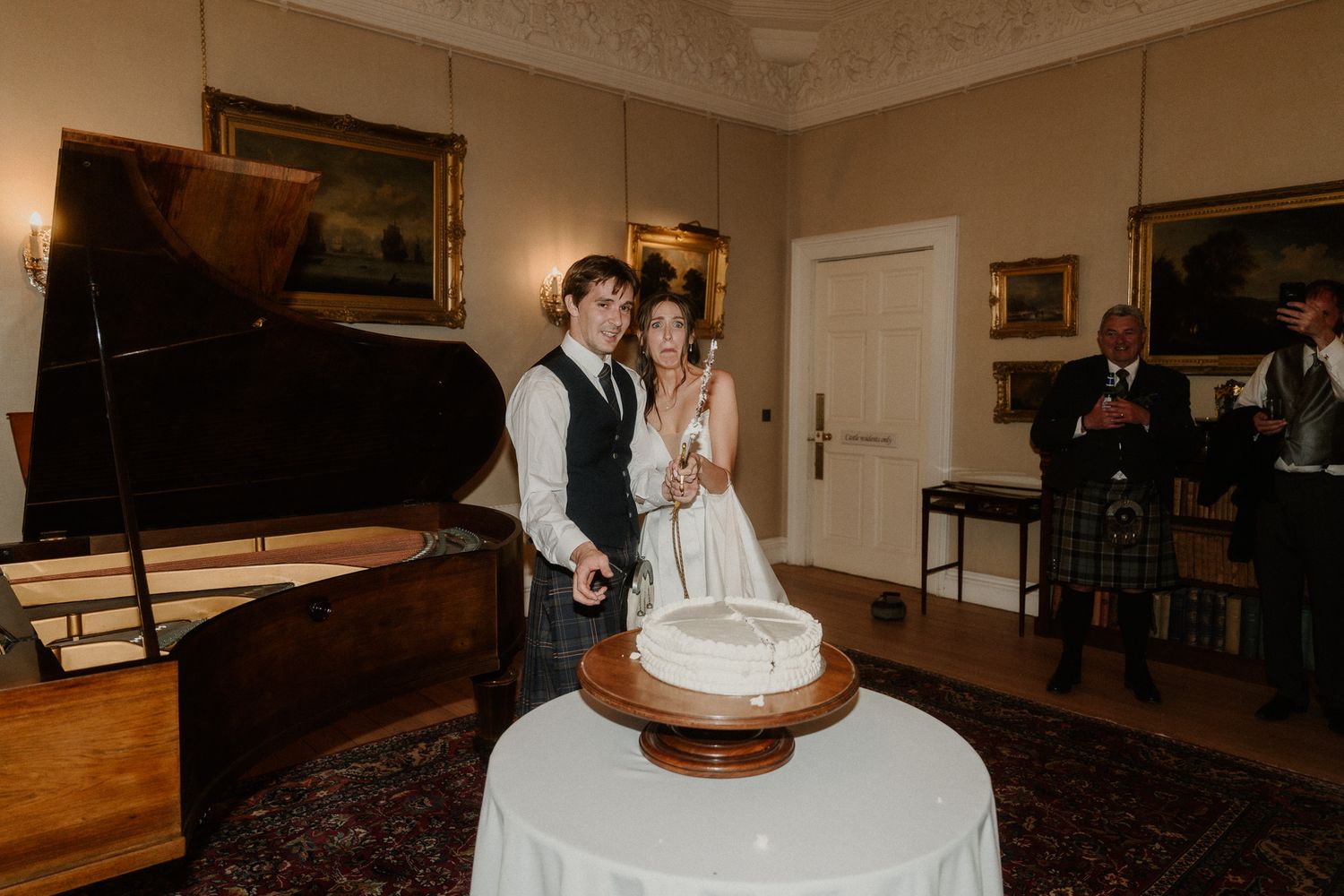 Bride and groom cutting white wedding cake in elegant room with grand piano, wearing formal attire.
