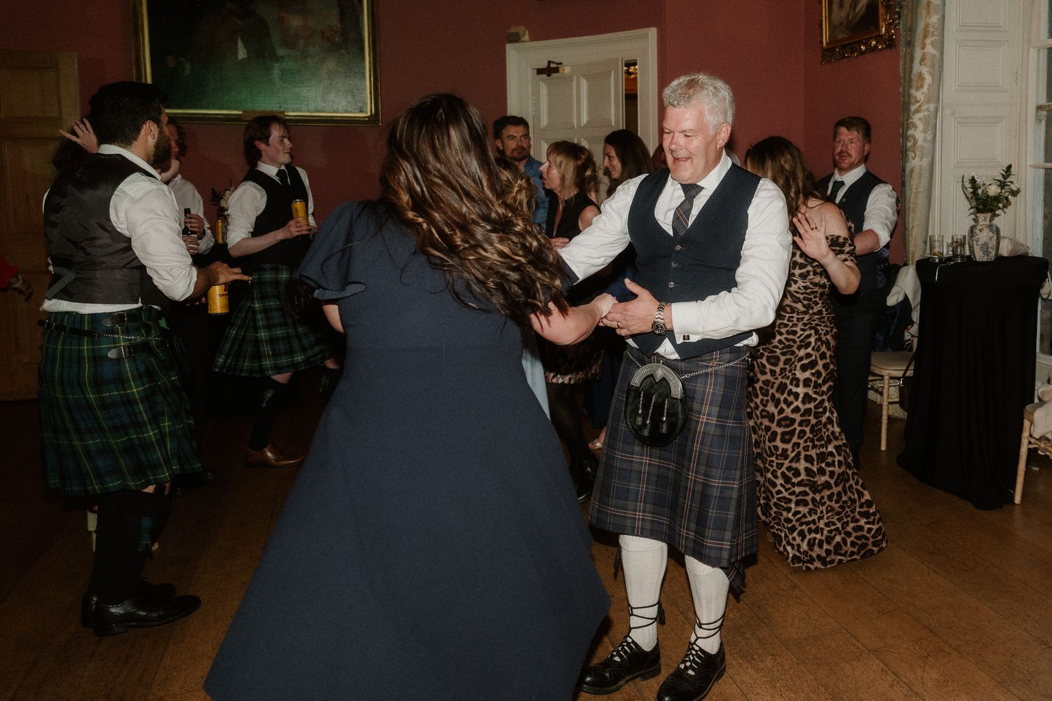 Guests in Scottish kilts and formal attire enjoy traditional ceilidh dancing at a wedding reception.