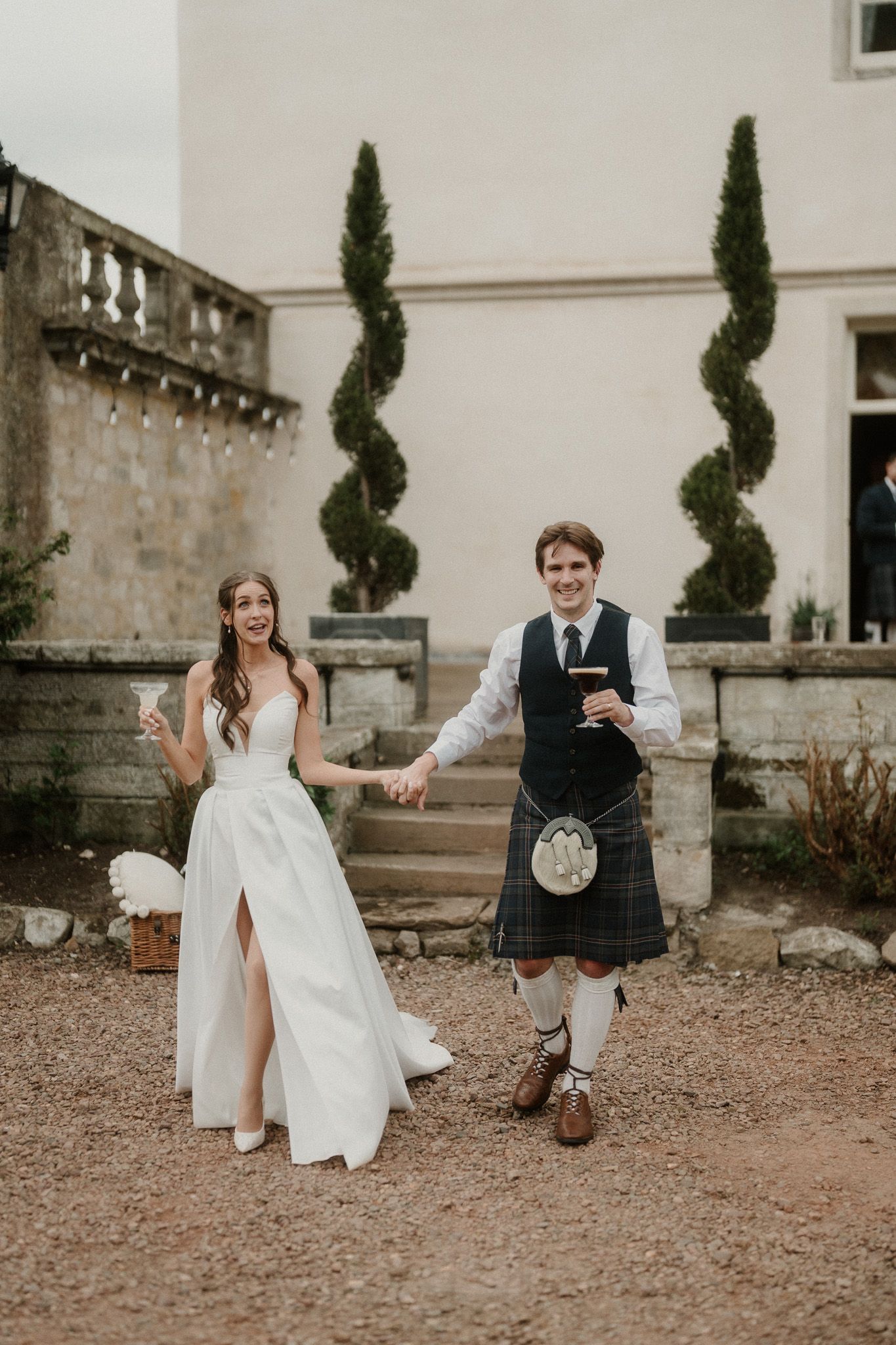 A joyful bride in a white gown and groom in a Scottish kilt hold hands outside a stone manor.