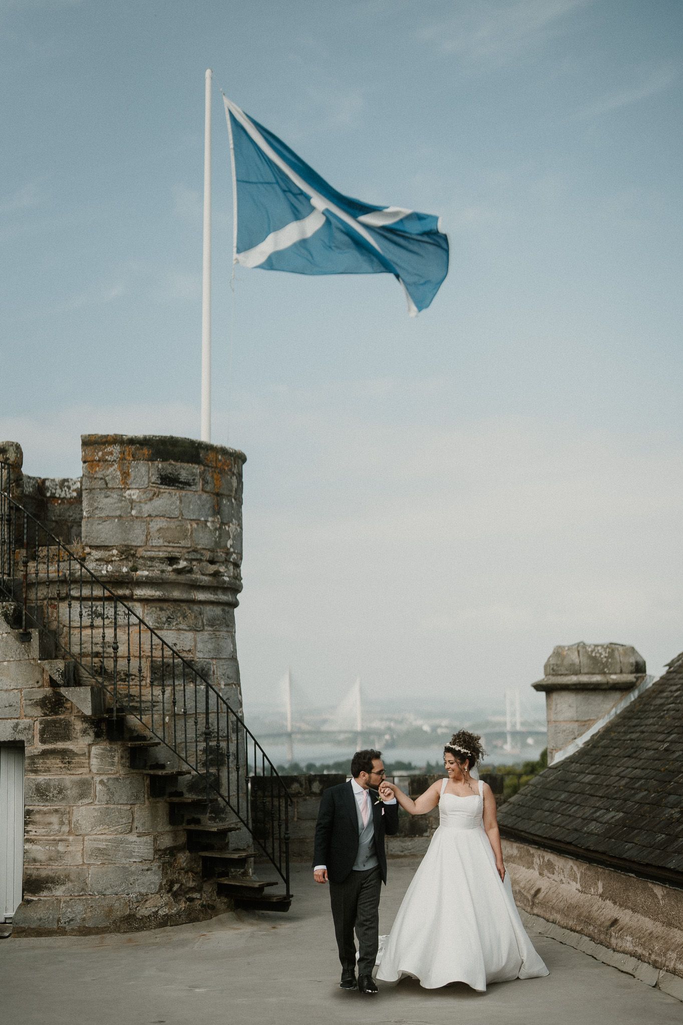Wedding couple walking along castle battlements with blue flag waving in the background.