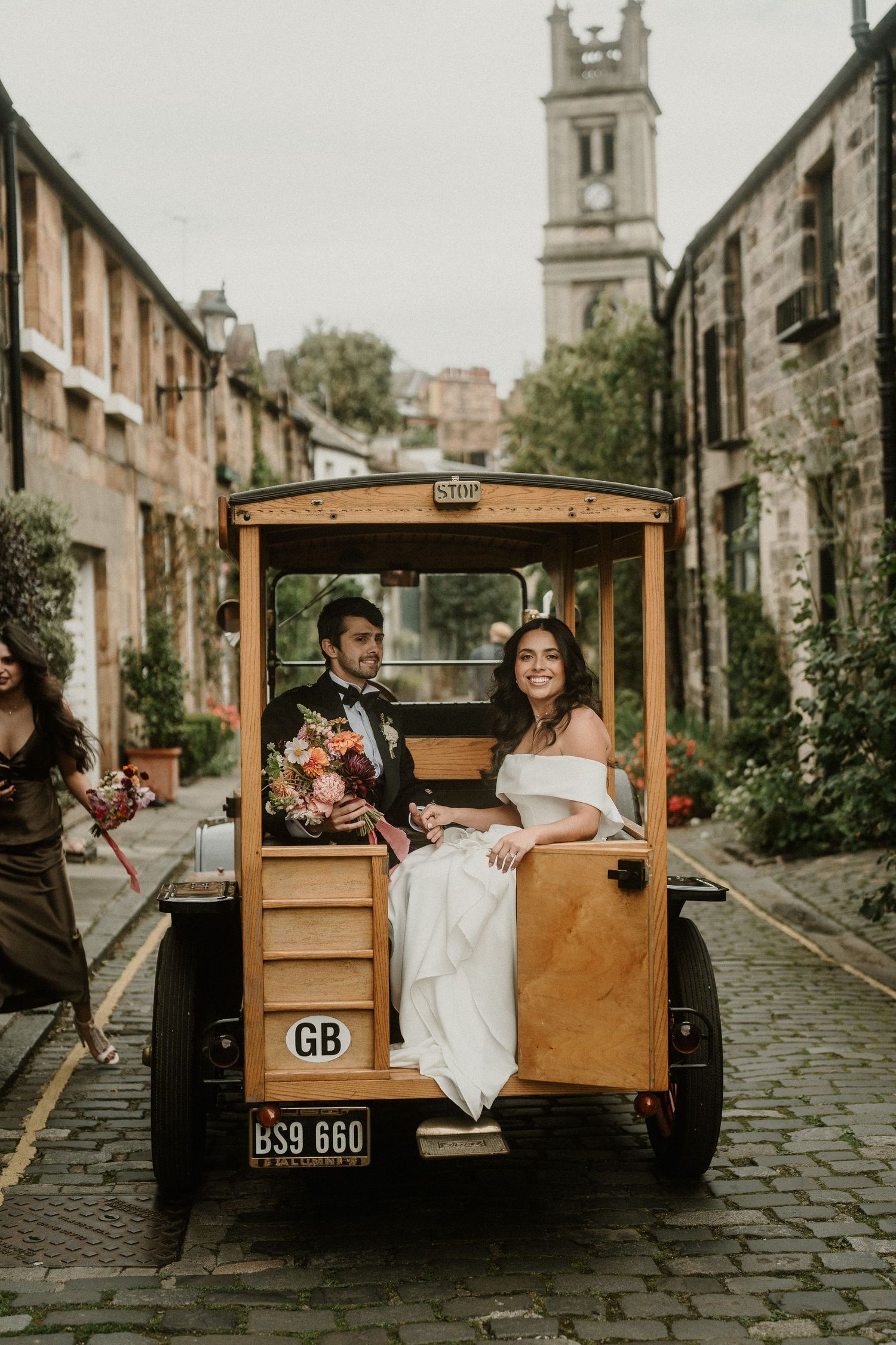 Couple riding in vintage wooden vehicle through historic stone-lined street with church spire in background.