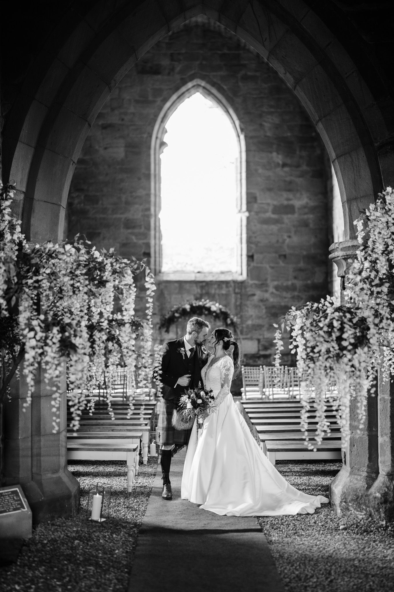 Romantic wedding scene in stone church undercroft with arched windows and floral decor.
