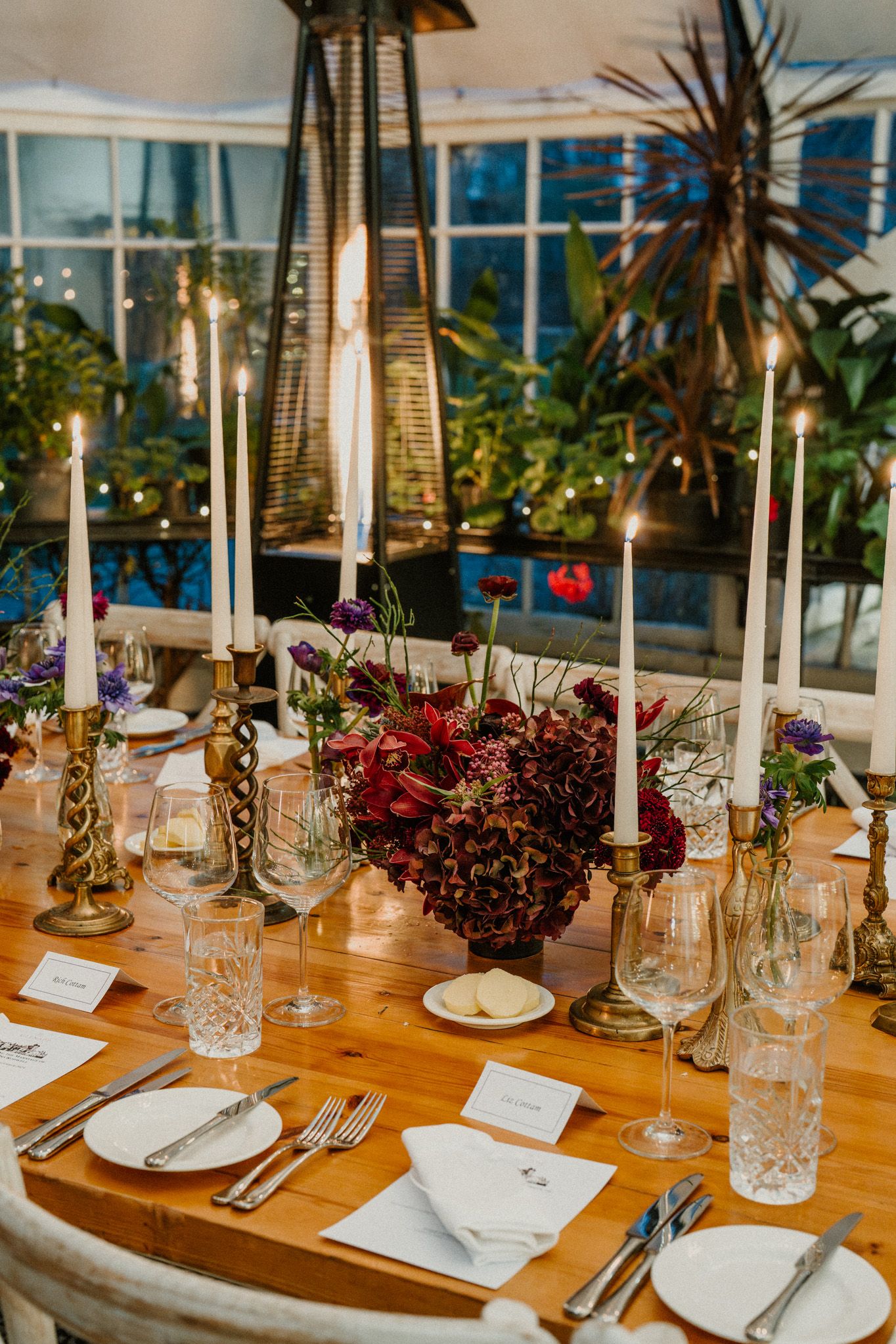Elegant wedding table setting with tall candles, crystal glassware, and deep red floral centerpieces in a greenhouse venue.